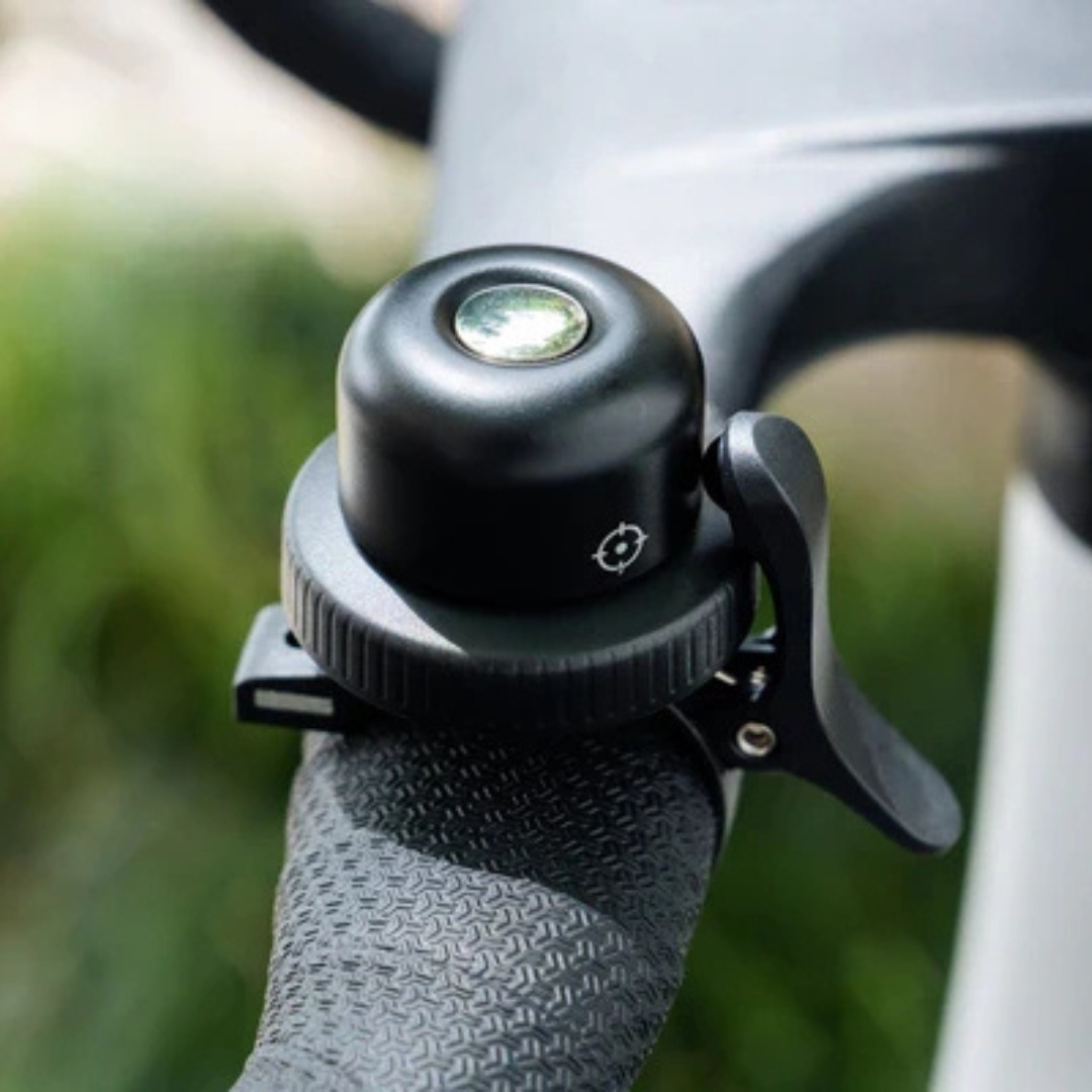 A black bicycle bell mounted on a textured handlebar with a blurred green background.