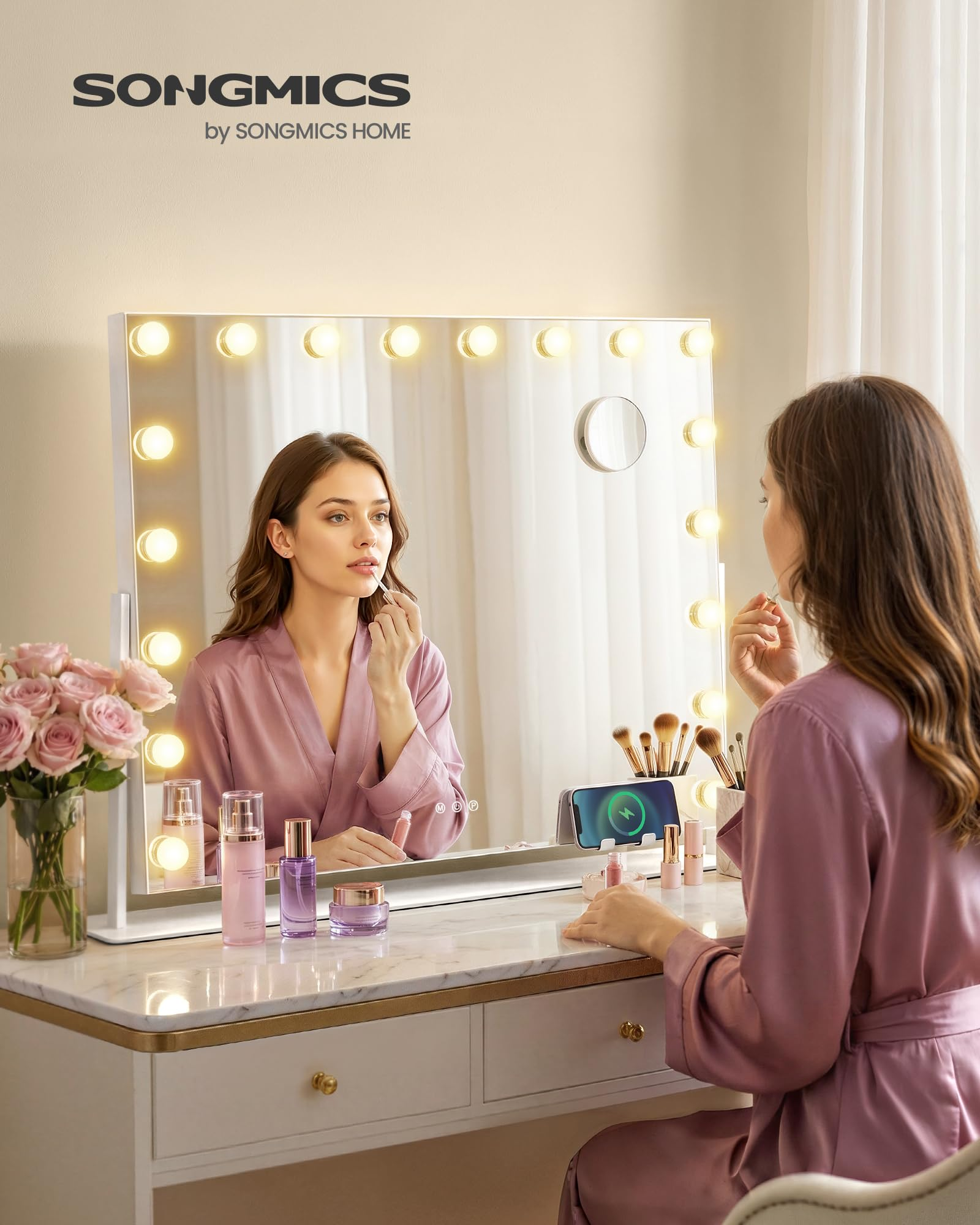 Una donna in vestaglia rosa applica il lucidalabbra davanti a uno specchio illuminato con cosmetici sul tavolo.