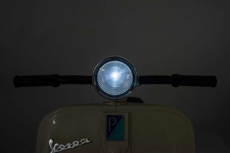 Ein Scheinwerfer eines beigen Vespa-Rollers leuchtet im Dunkeln.
