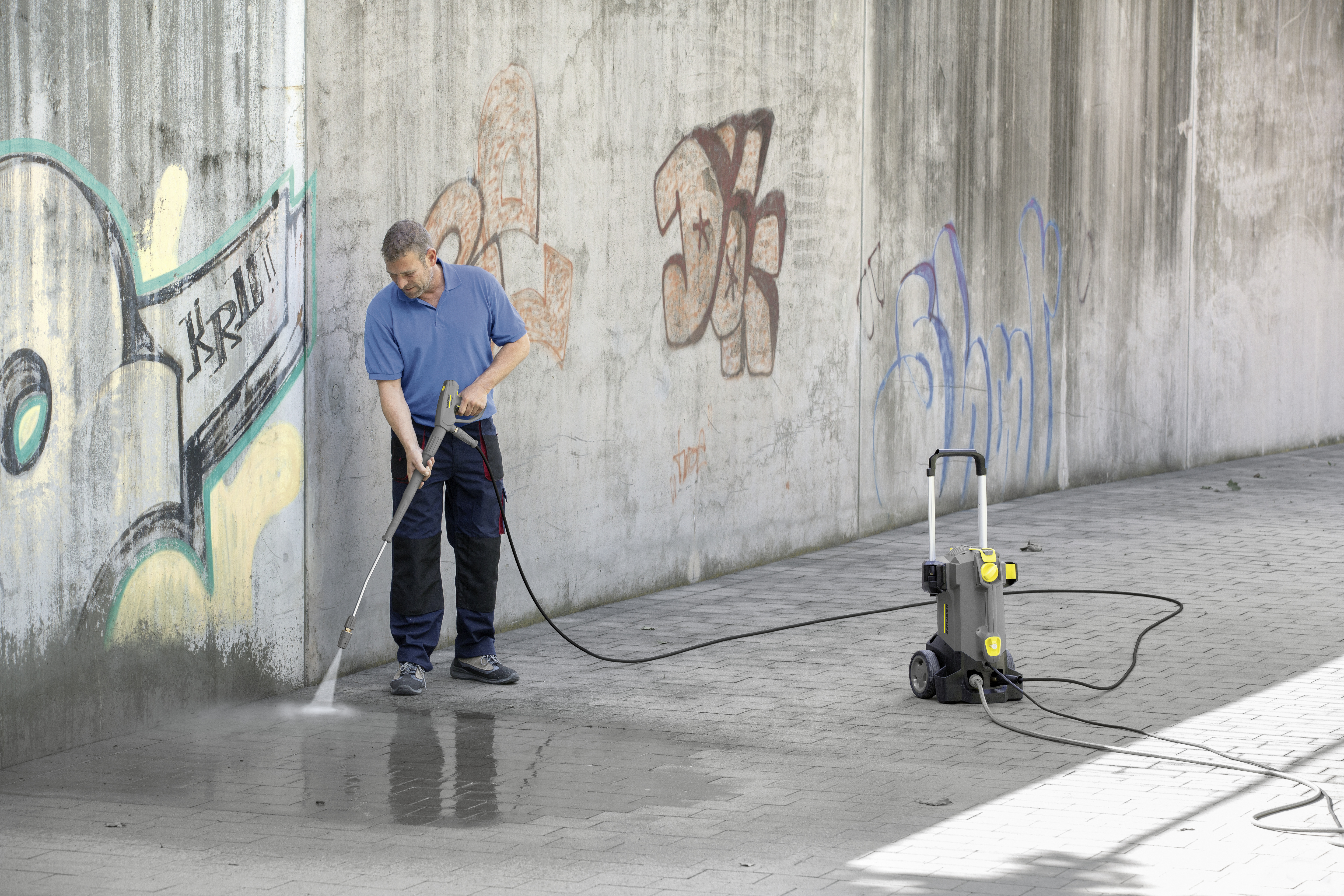 Ein Mann in blauer Kleidung reinigt eine graffiti-verschmierte Betonwand mit einem Hochdruckreiniger.