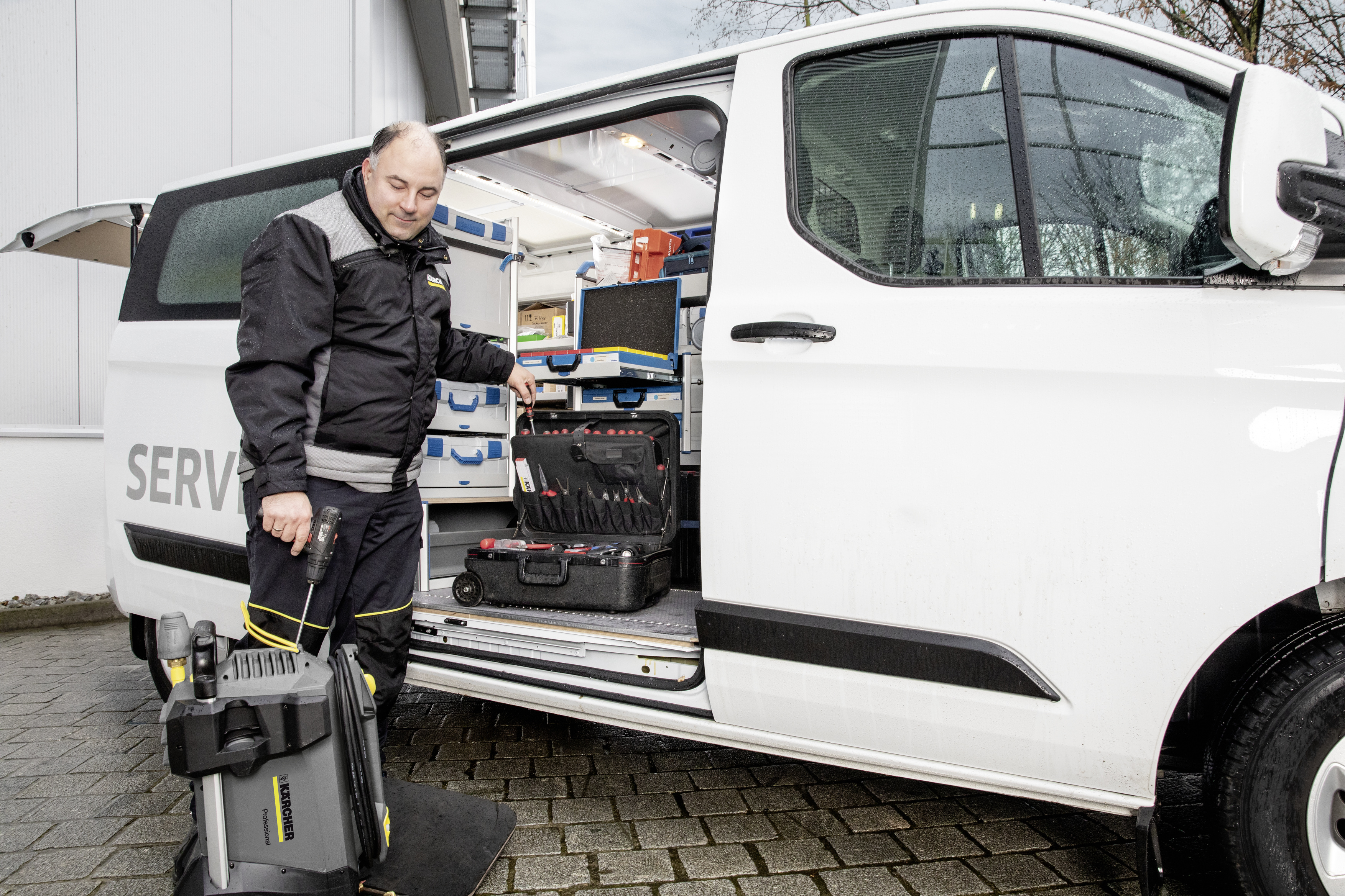 Ein Techniker steht neben einem weißen Service-Van mit geöffneter Tür und hält Werkzeug und ein Reinigungsgerät.