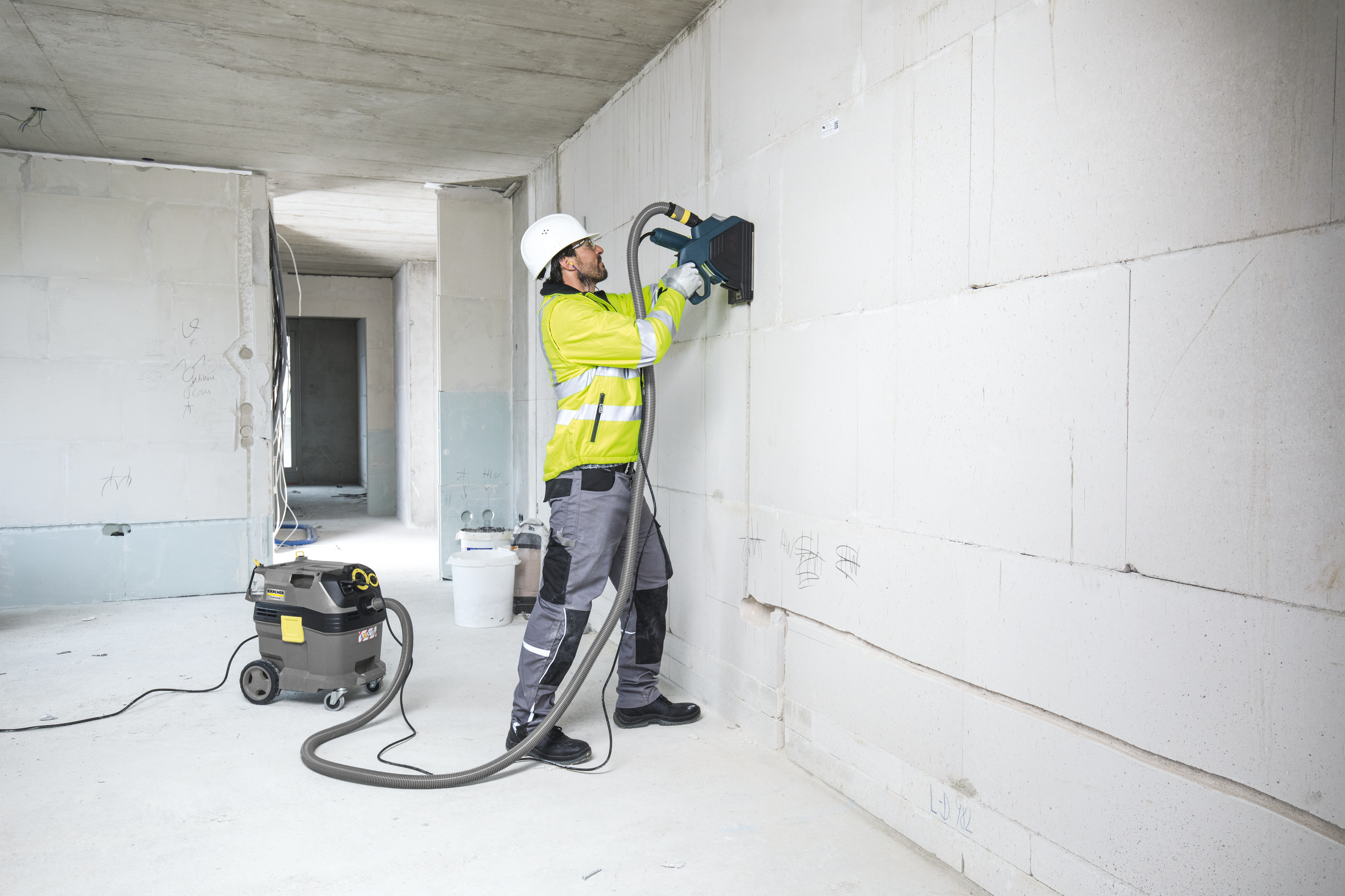 Ein Arbeiter mit weißem Helm und gelber Jacke benutzt ein Elektrowerkzeug an einer Betonwand.