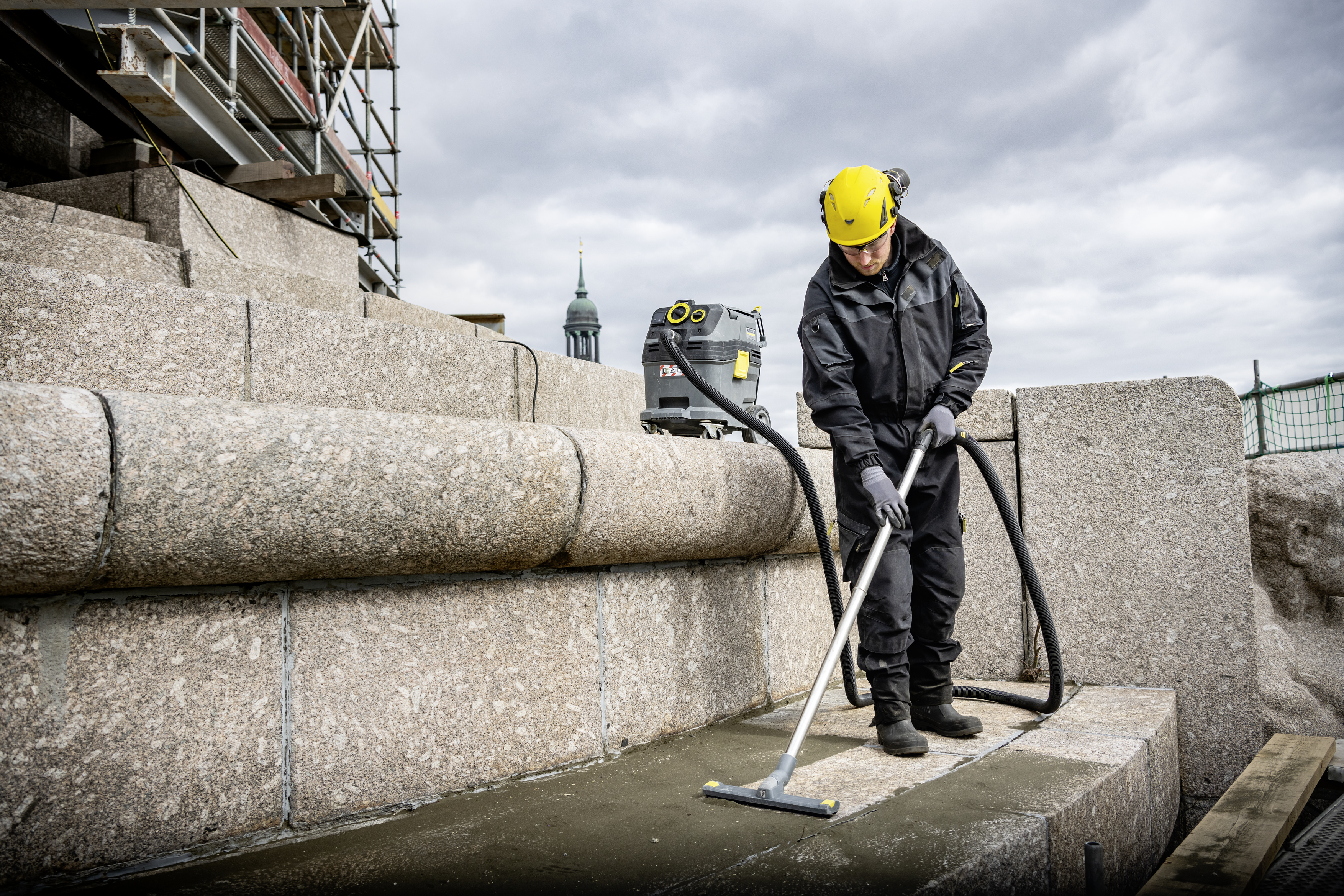 Ein Arbeiter mit gelbem Helm reinigt Steinstufen mit einem großen Industriestaubsauger.