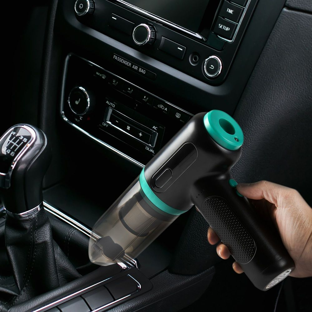 Una mano tiene un aspirapolvere portatile compatto nero e verde acqua nella console centrale di un'auto.