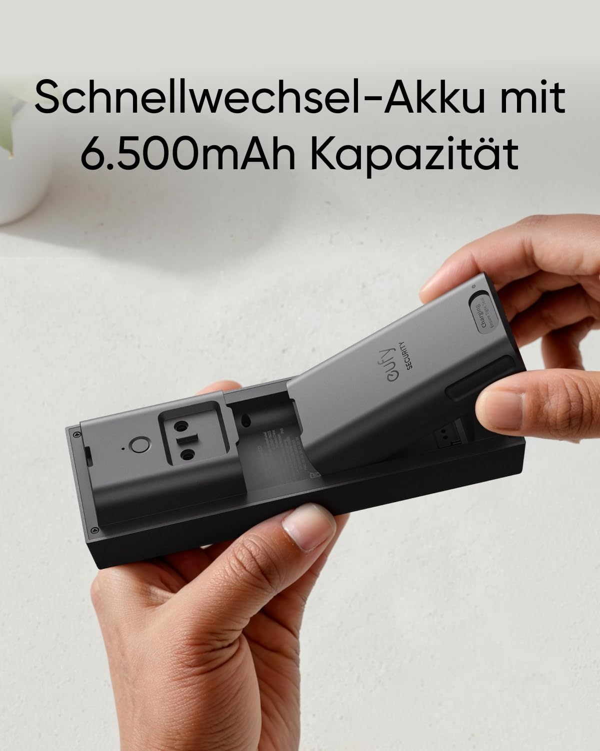 Hände legen einen grauen Akku in ein Eufy-Sicherheitskamera-Gehäuse auf weißem Hintergrund.