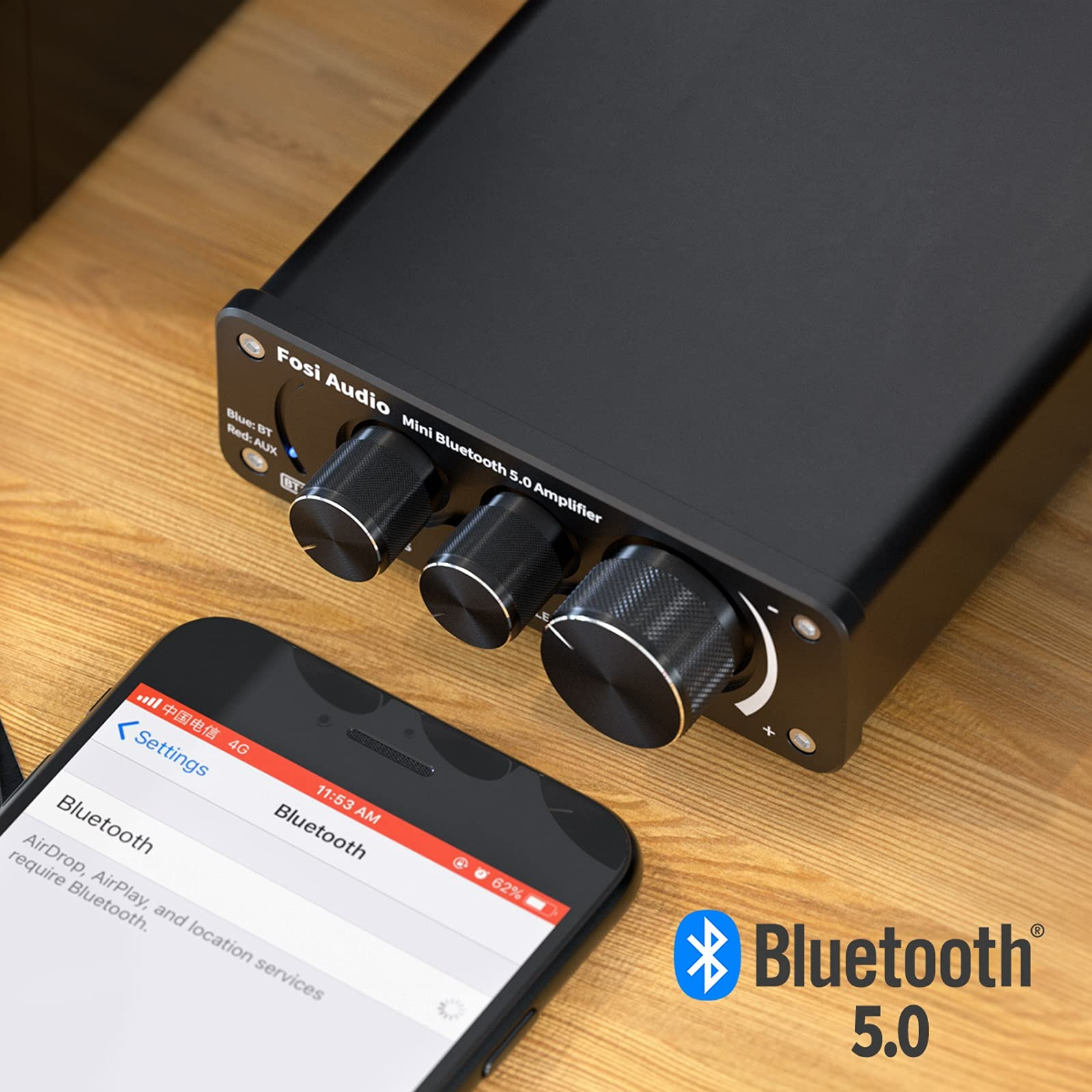 Ein schwarzer Fosi Audio Mini Bluetooth 5.0 Verstärker neben einem Smartphone mit Bluetooth-Einstellungen auf Holz.