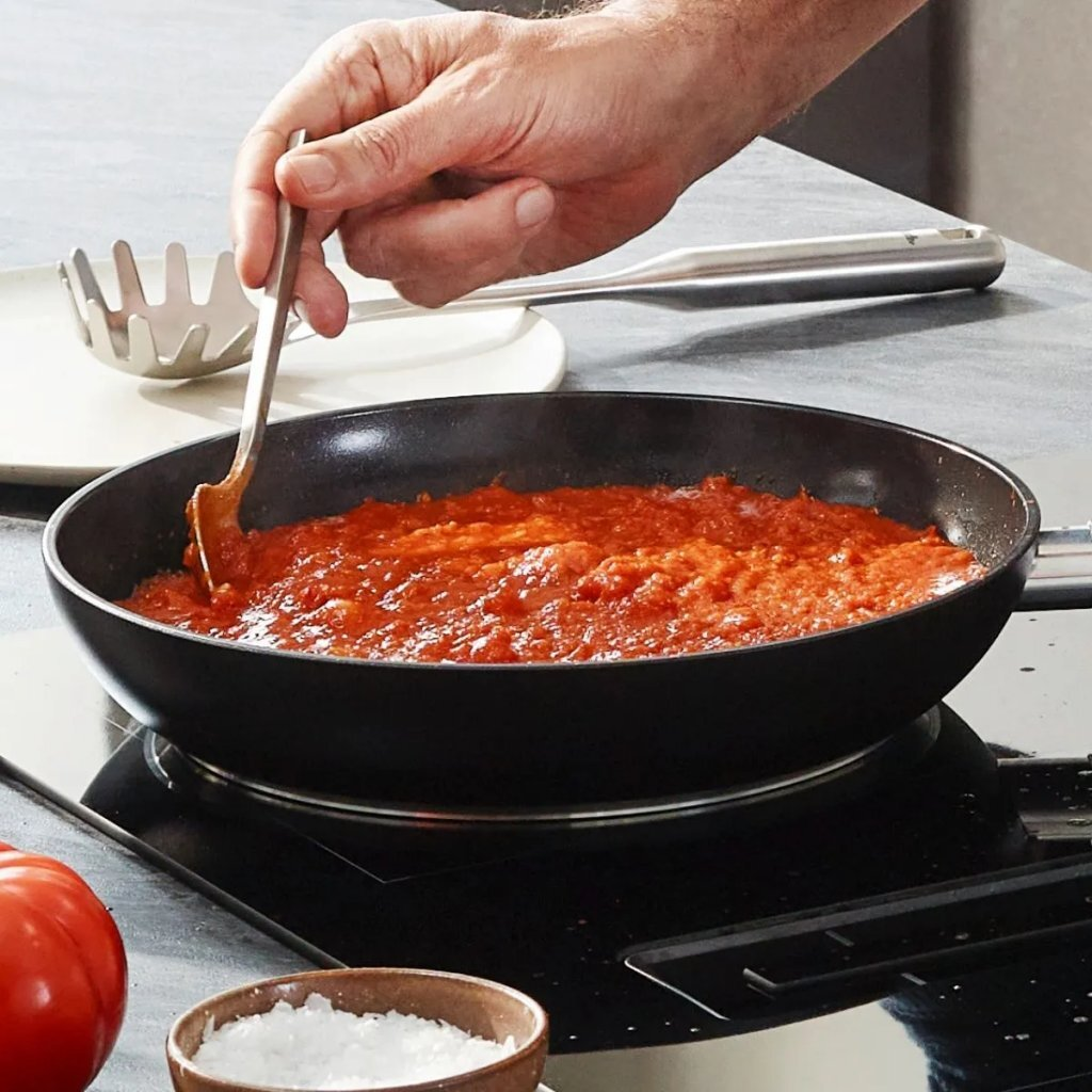 Une main remue de la sauce tomate rouge dans une poêle noire sur une cuisinière.