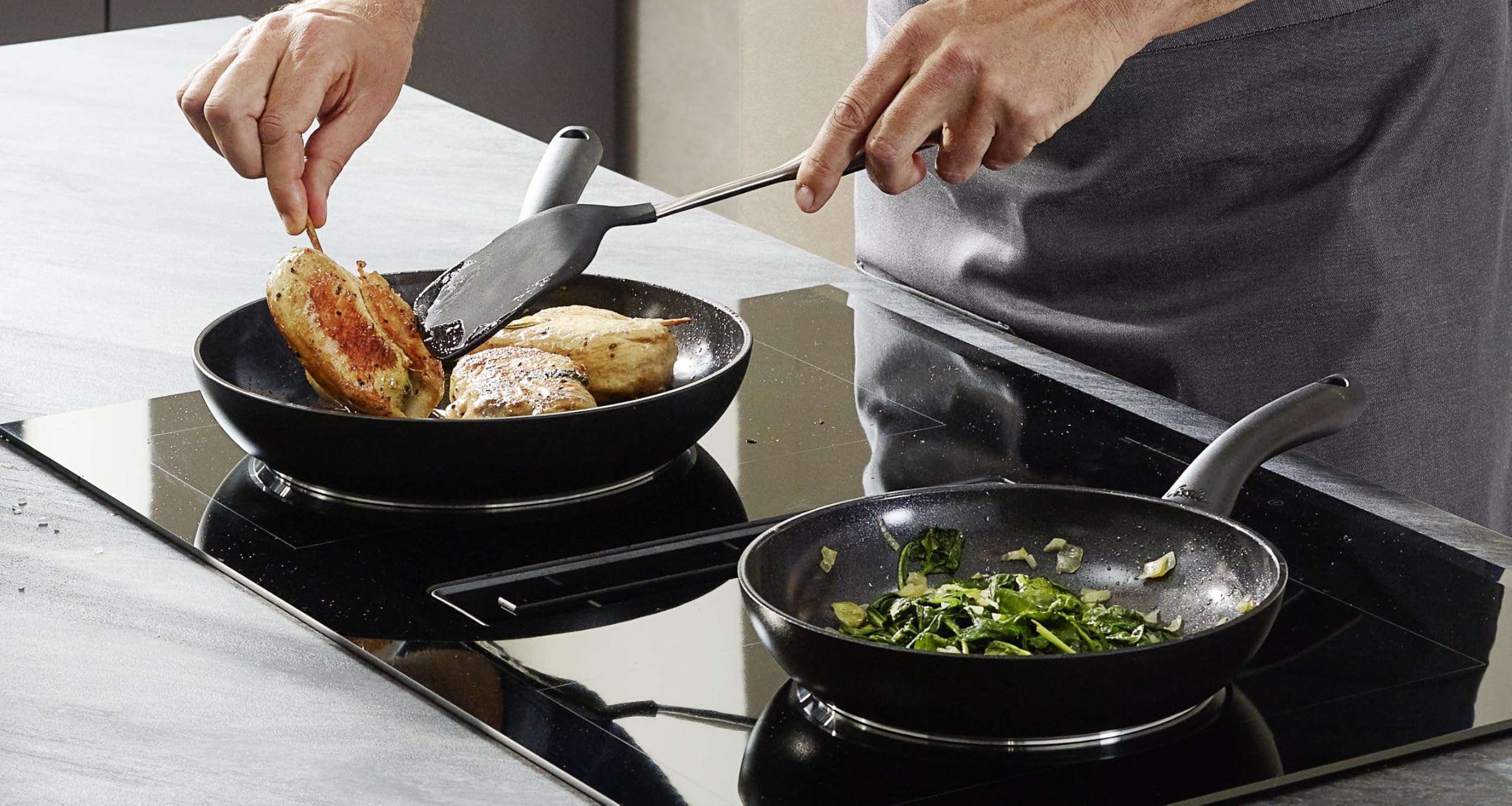 Un cuisinier fait frire des blancs de poulet dans une poêle et des épinards dans une autre sur une plaque.