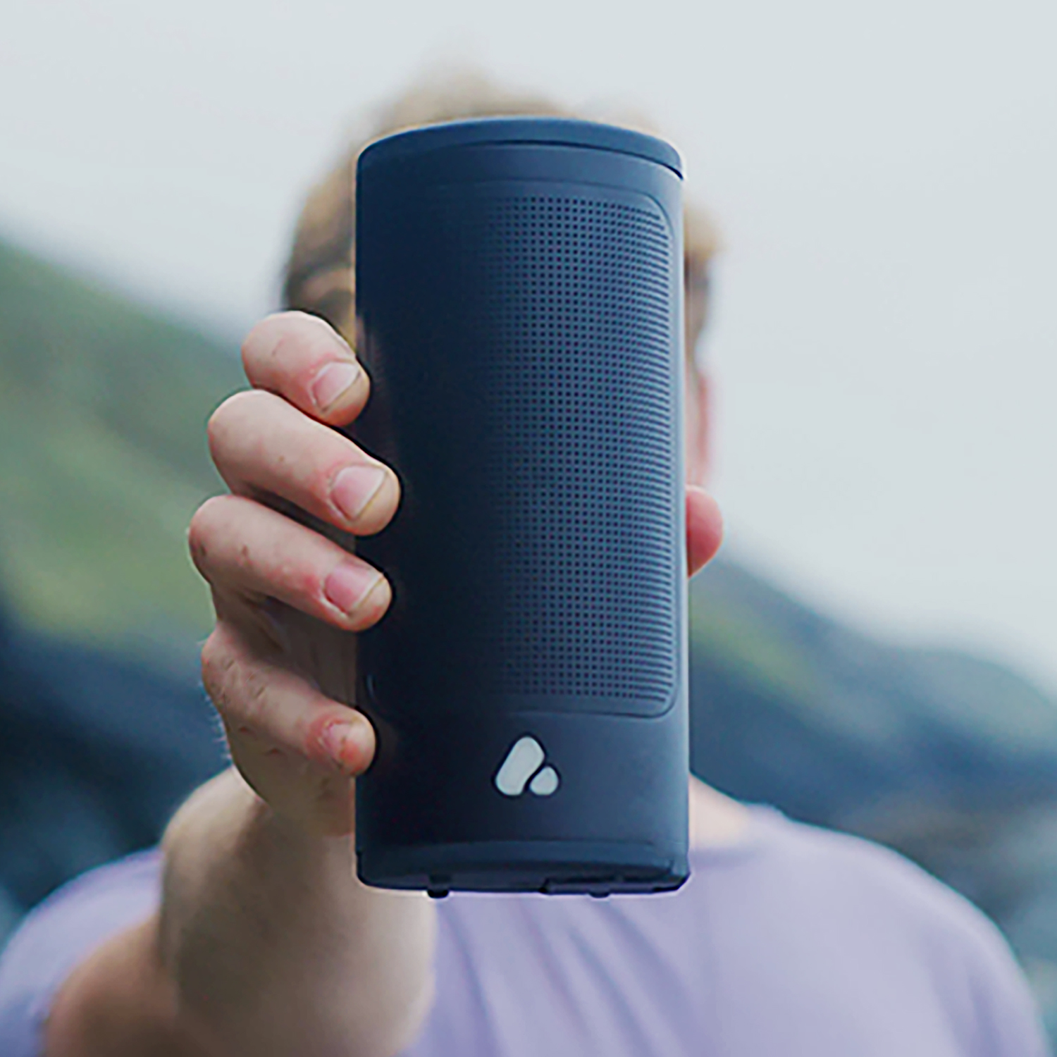 Une personne tient une enceinte Bluetooth cylindrique sombre avec un petit logo blanc, en extérieur.