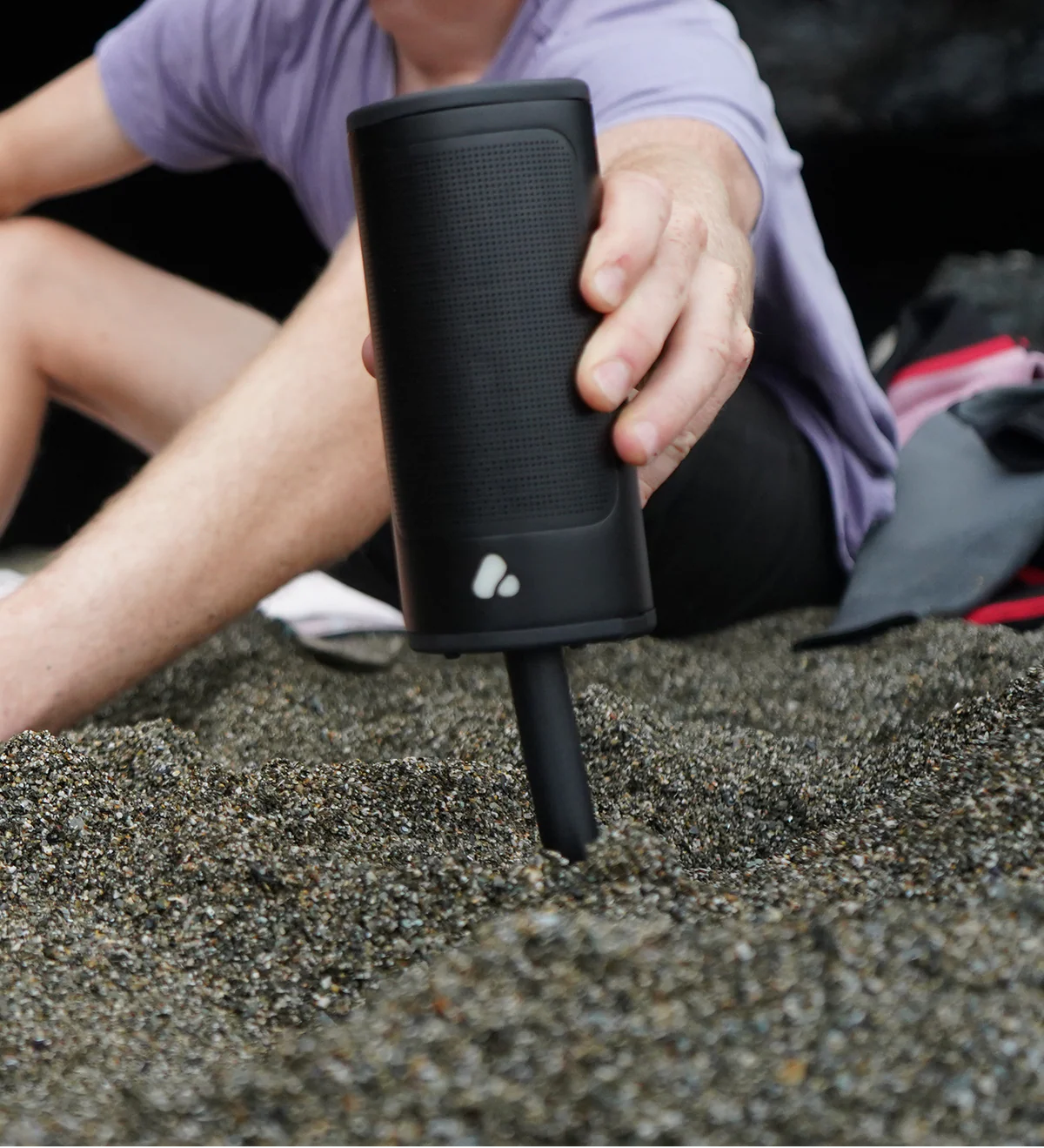 Une personne enfonce un haut-parleur cylindrique noir avec logo dans le sable d'une plage.