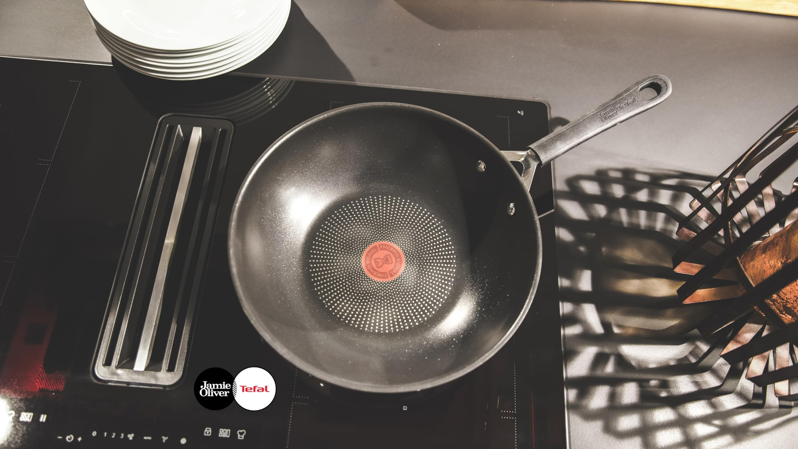 Eine schwarze Jamie Oliver Tefal Wok-Pfanne auf einem Induktionskochfeld mit gestapelten Tellern im Hintergrund.