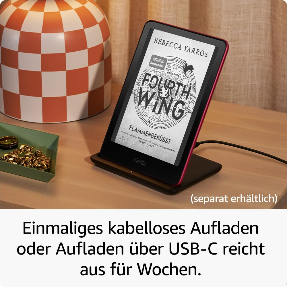 Une liseuse Kindle affichant le livre Fourth Wing sur un support de charge sur une table en bois, à côté d'une lampe.