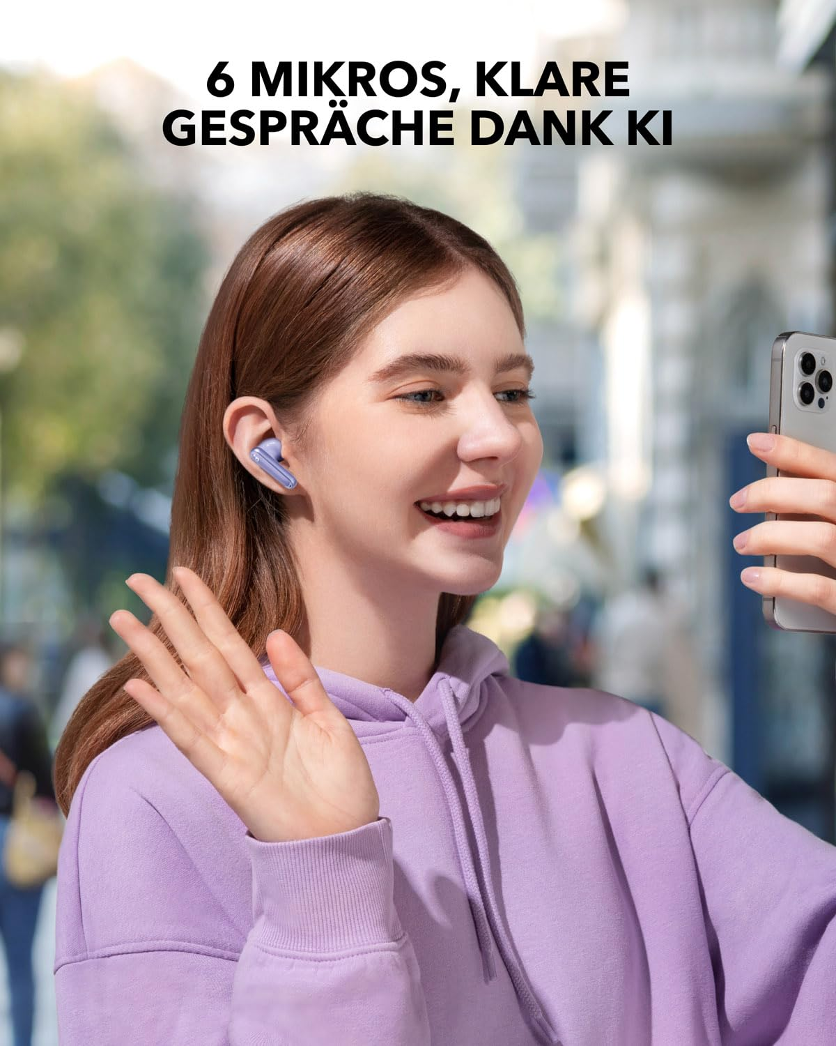 Une femme souriante en sweat à capuche violet tient un smartphone, fait signe et porte des écouteurs.