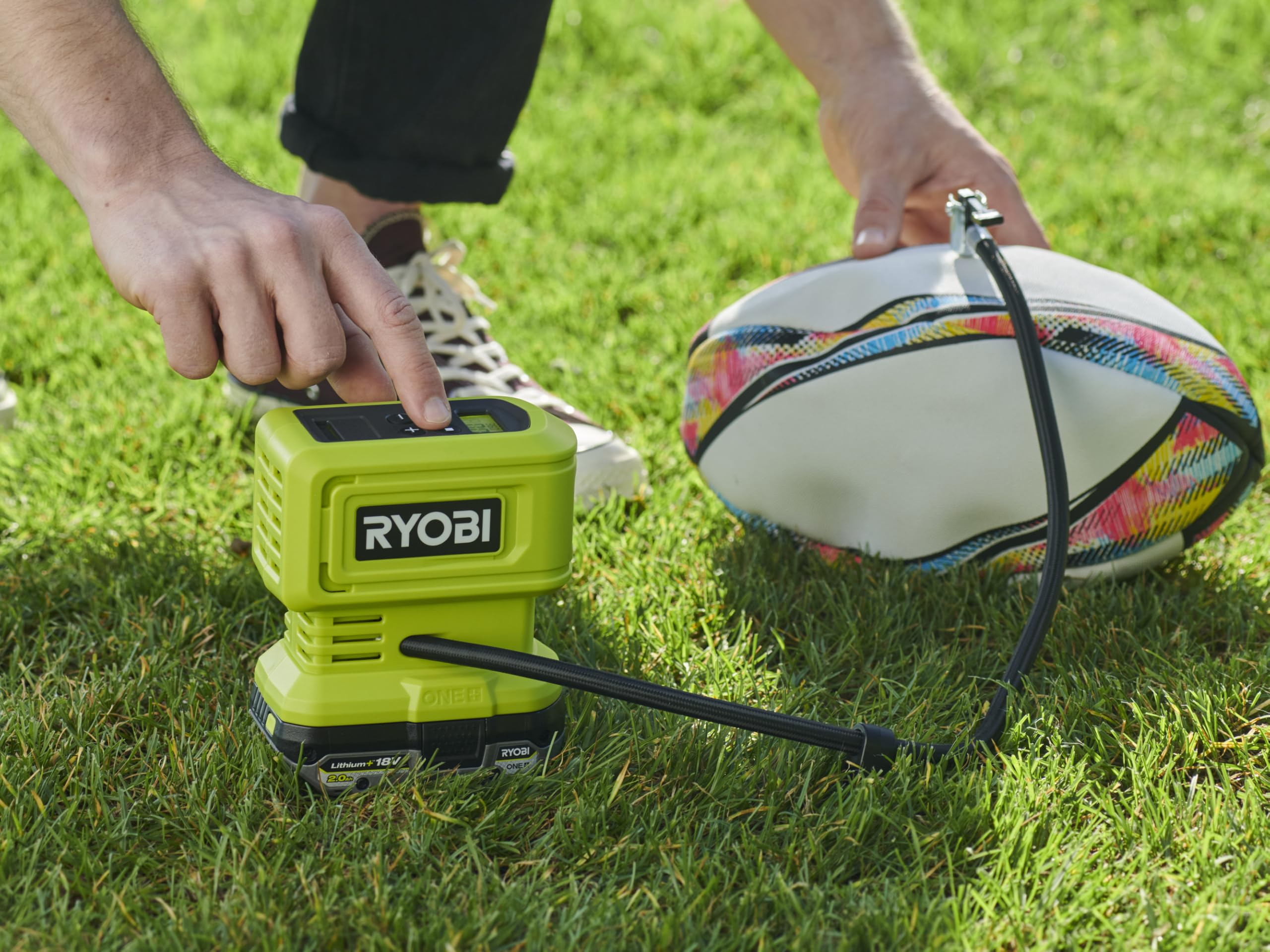 Person bedient eine grüne Ryobi-Luftpumpe auf Rasen neben einem bunten Rugbyball.