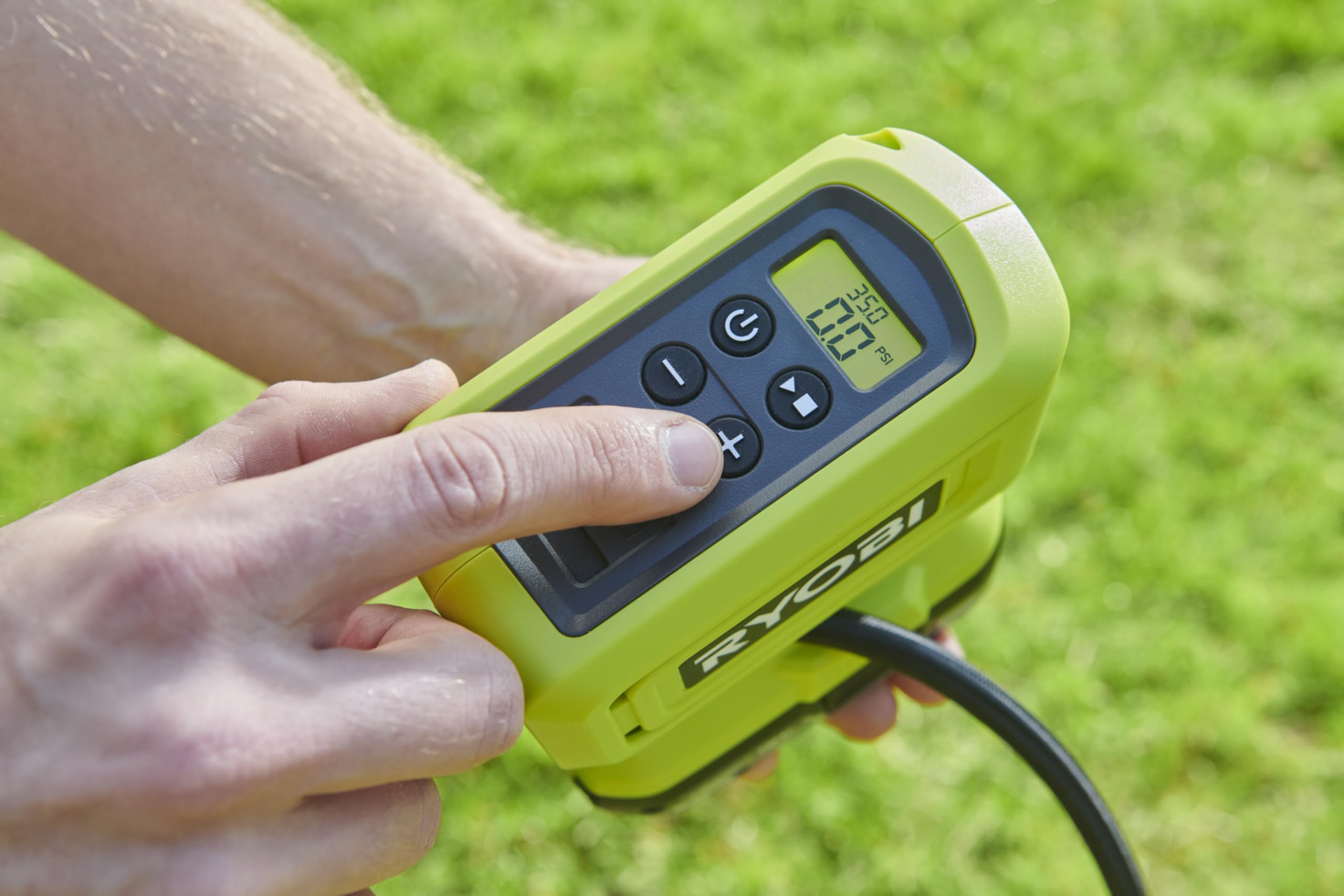 Eine Person hält einen grünen digitalen Ryobi-Luftkompressor und drückt Tasten vor grasgrünem Hintergrund.