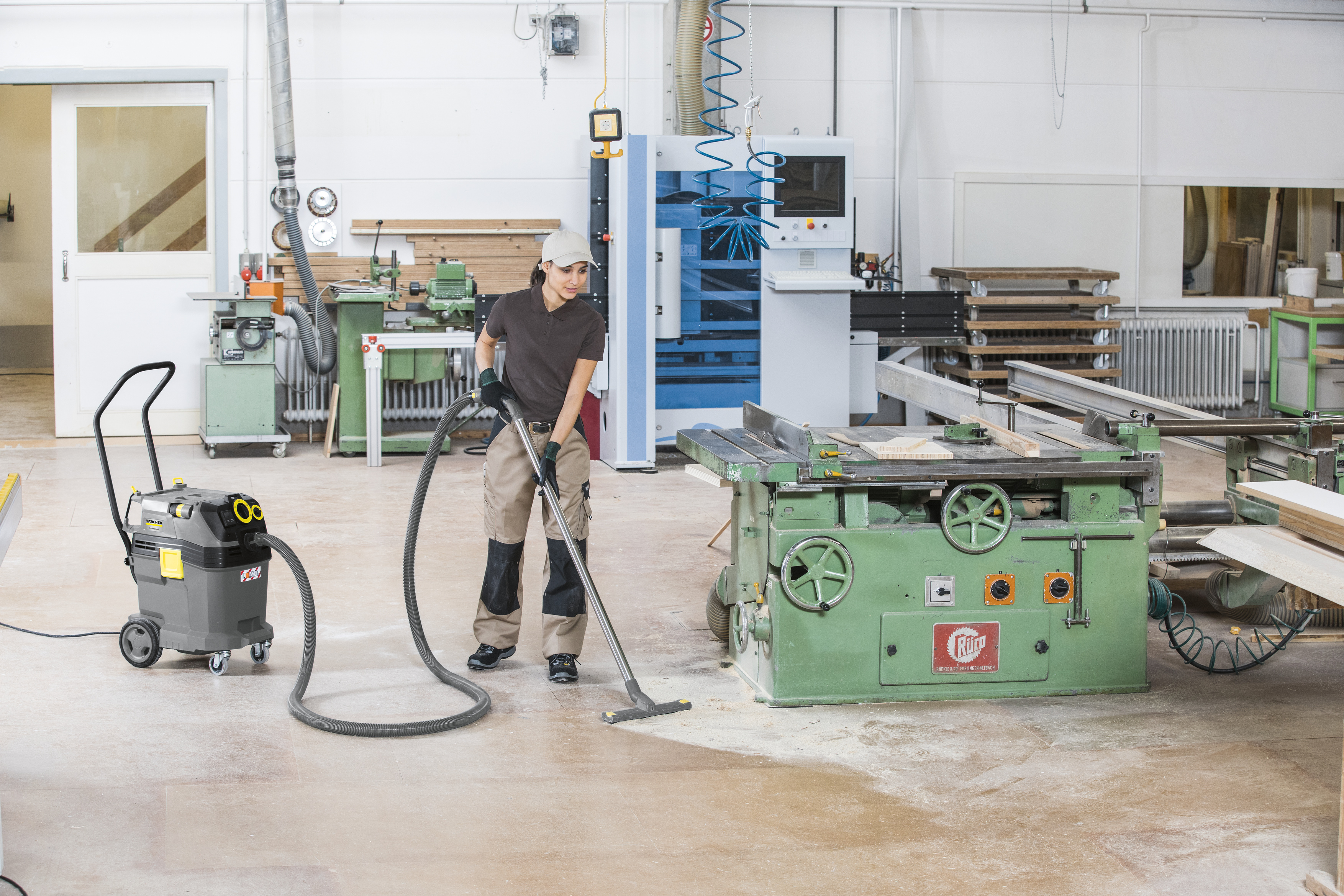 Eine Arbeiterin reinigt mit einem Industriesauger den Boden einer Werkstatt neben großen Maschinen.