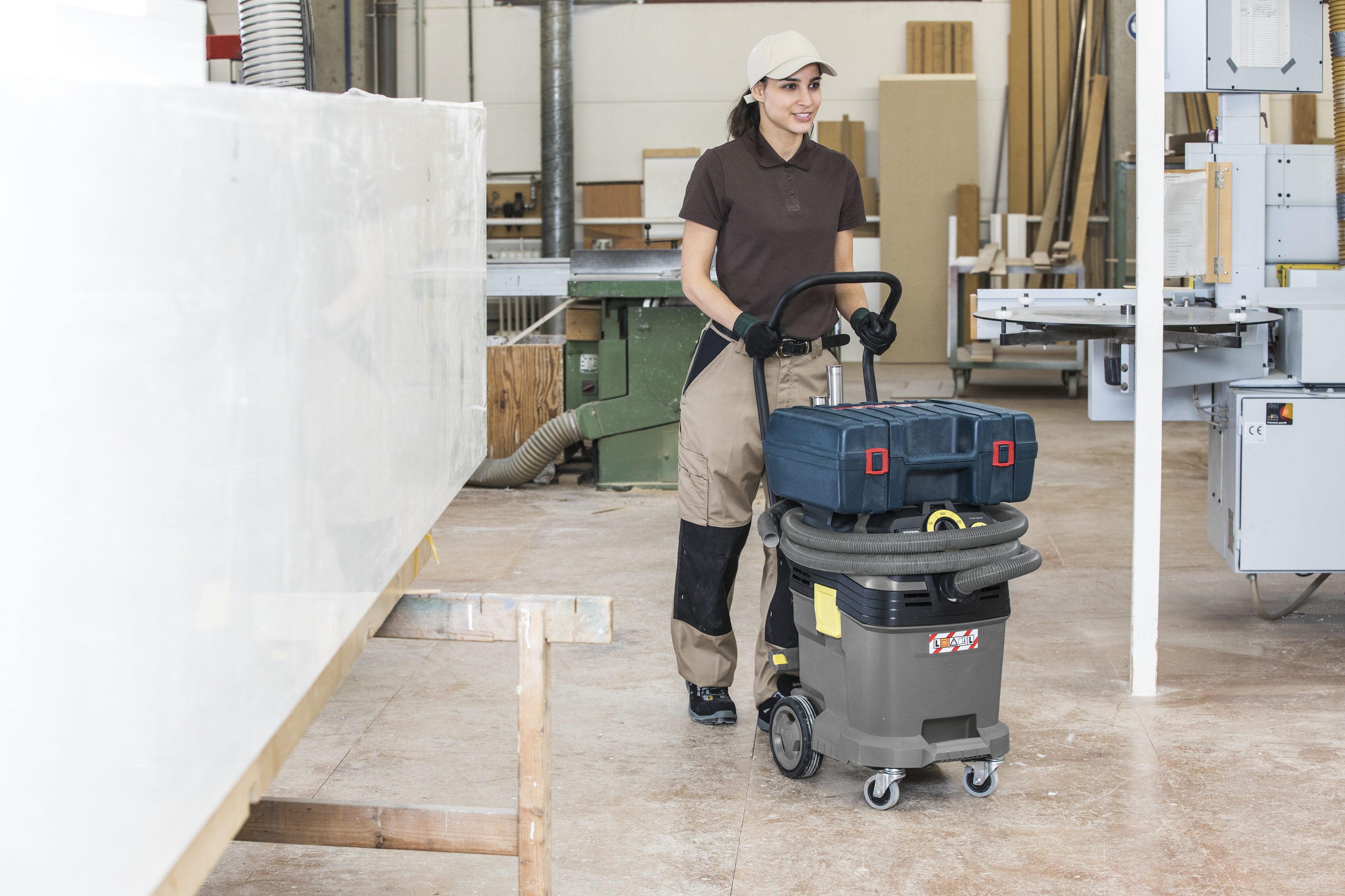 Eine Handwerkerin schiebt einen Werkstattstaubsauger mit Werkzeugkasten in einer Werkstatt.