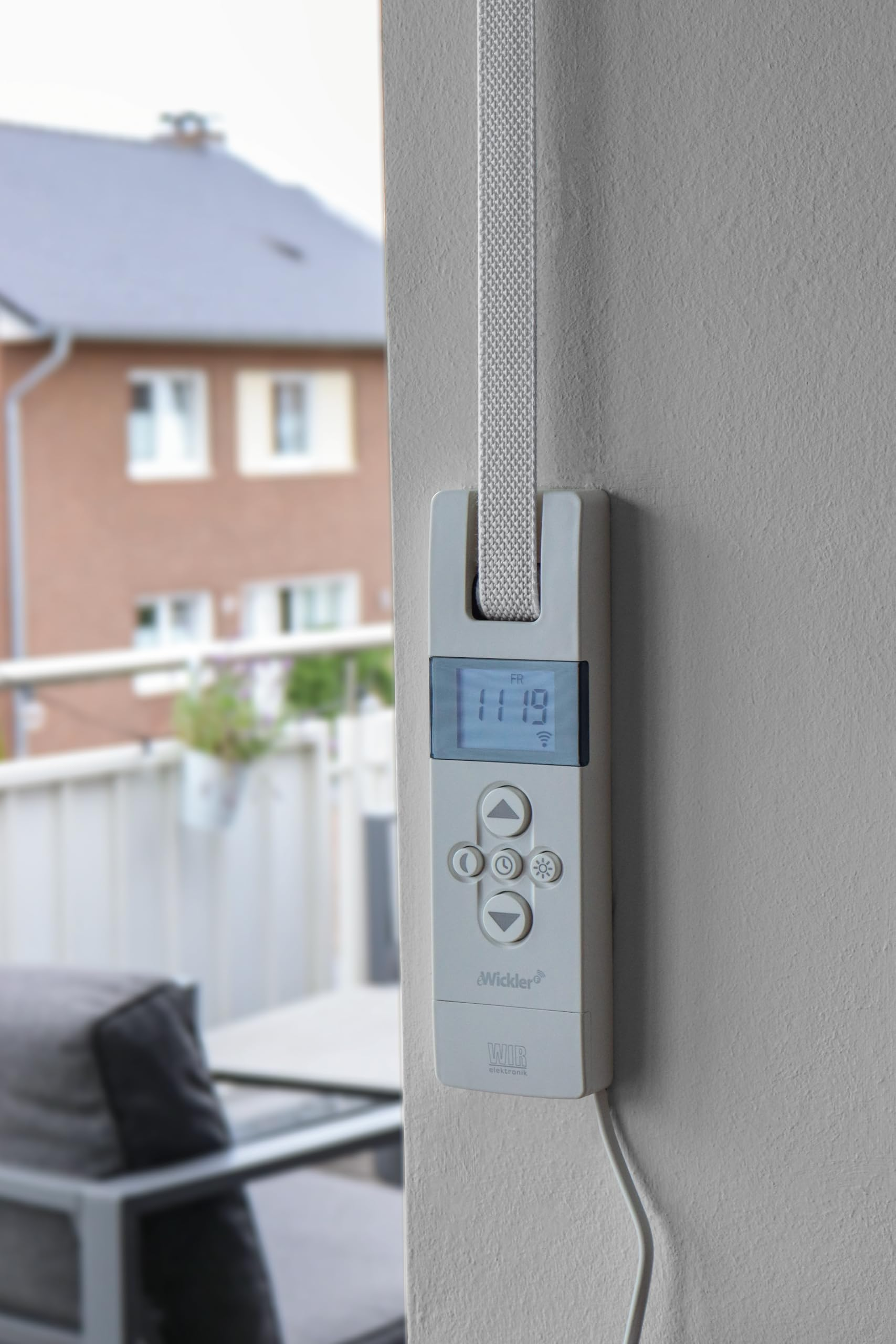 Ein weißer elektronischer Gurtwickler mit Display und Tasten an einer Wand, im Hintergrund ein Gebäude.
