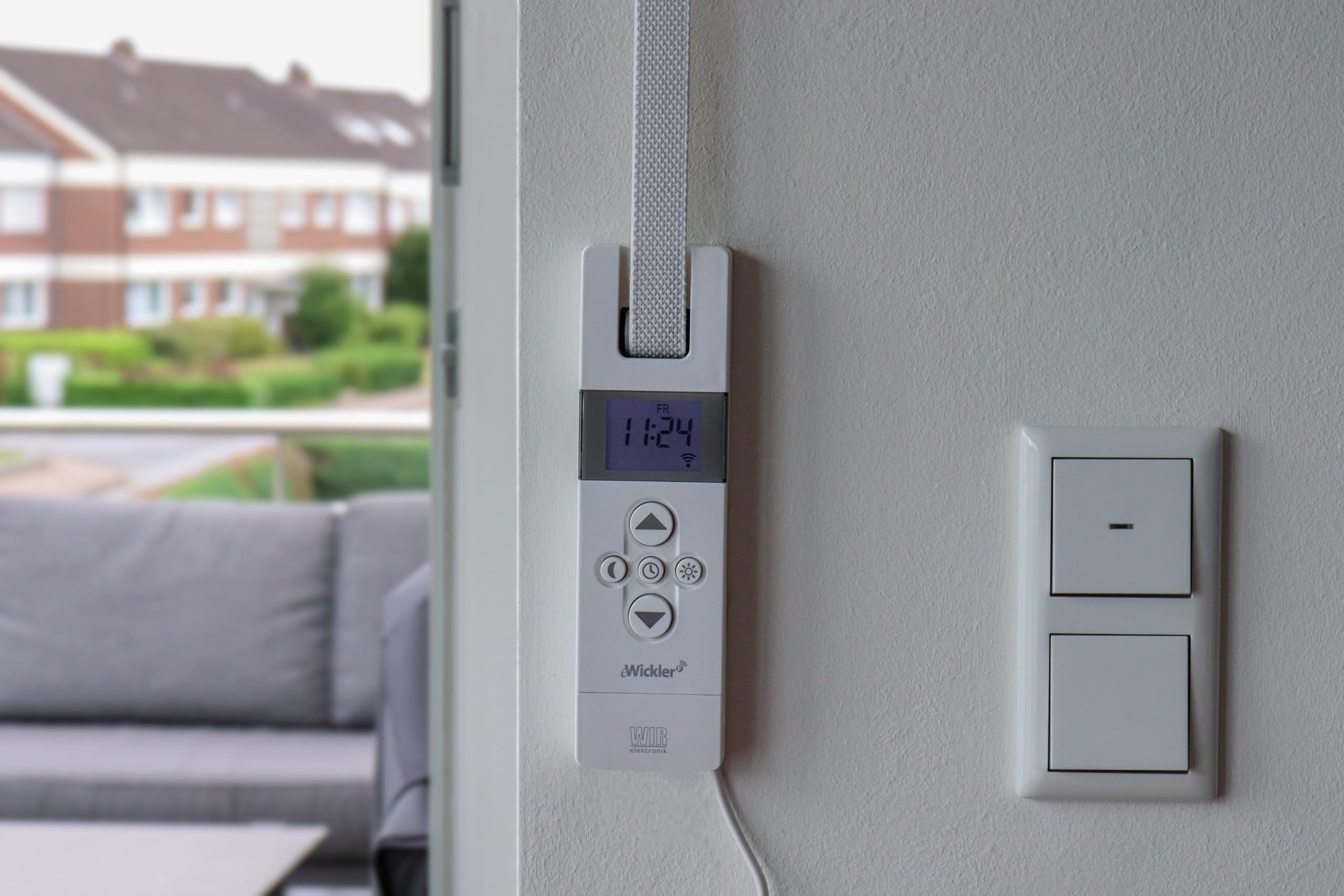 Ein weißer elektronischer Rollladenbedienteil an der Wand neben einem Schalter, mit Blick auf Häuser draußen.