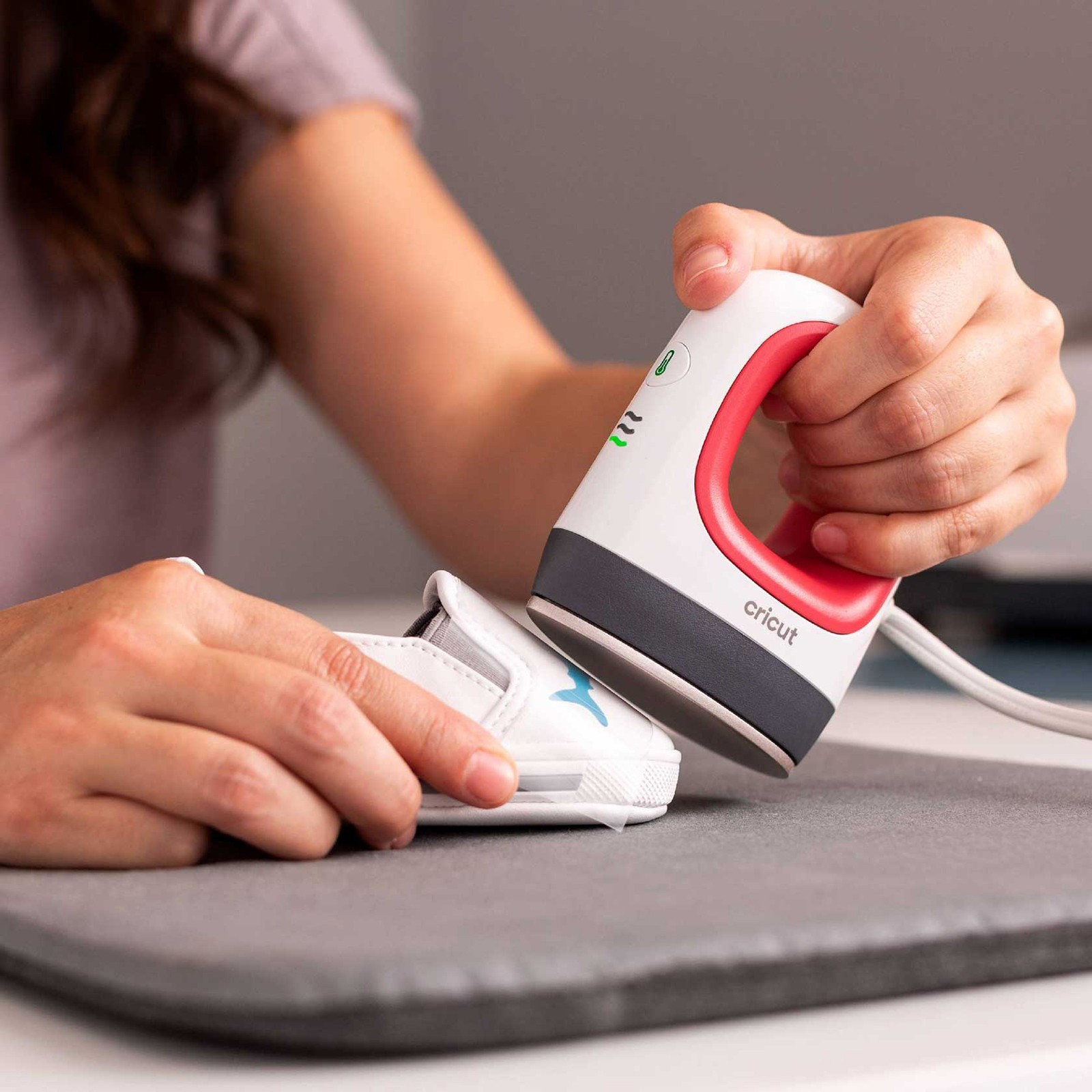 Eine Person benutzt eine kompakte weiße und rote Cricut-Heißpresse auf einem kleinen weißen Schuh auf einer grauen Matte.