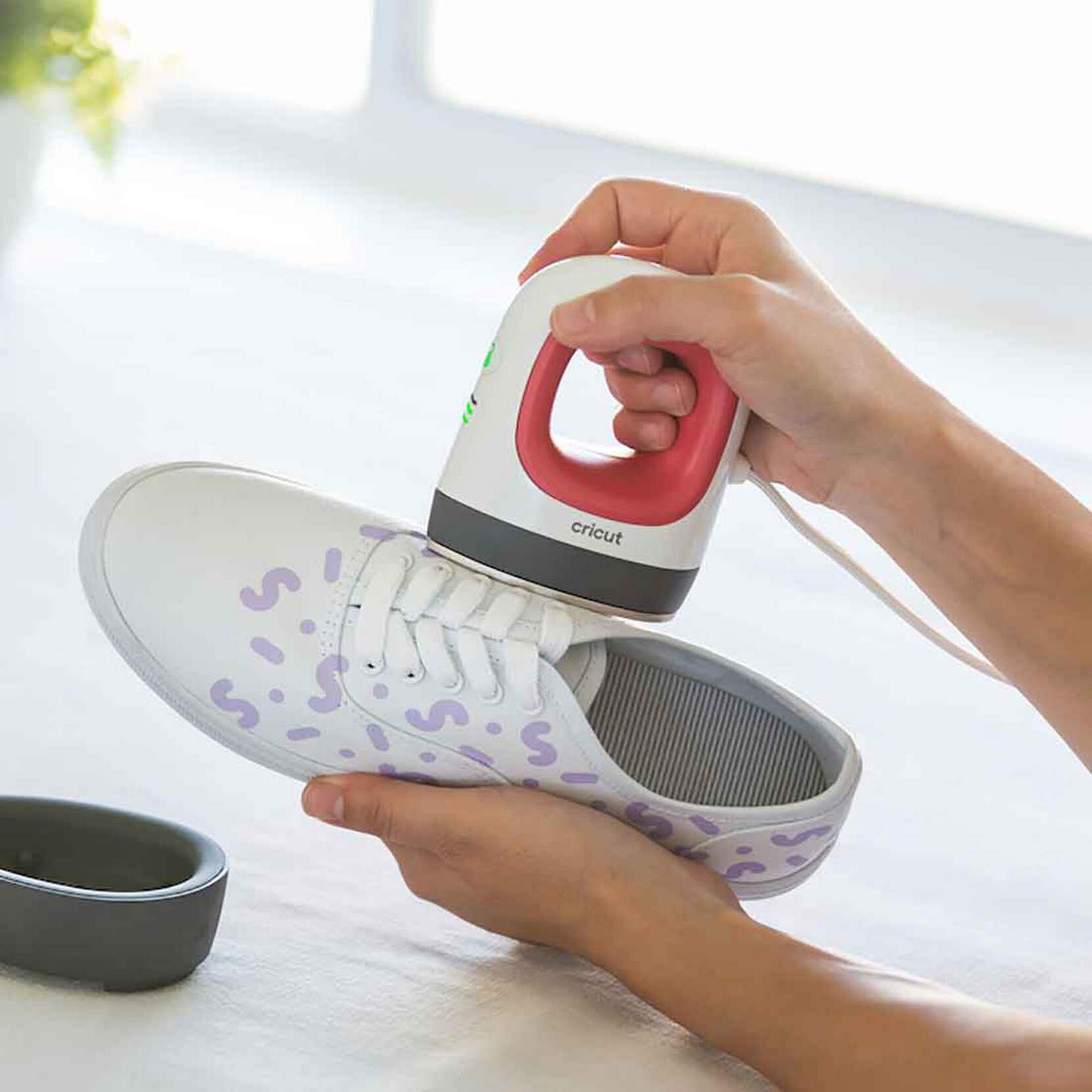 Eine Hand benutzt eine Cricut-Mini-Bügelpresse auf einem weißen Schuh mit lila Mustern.