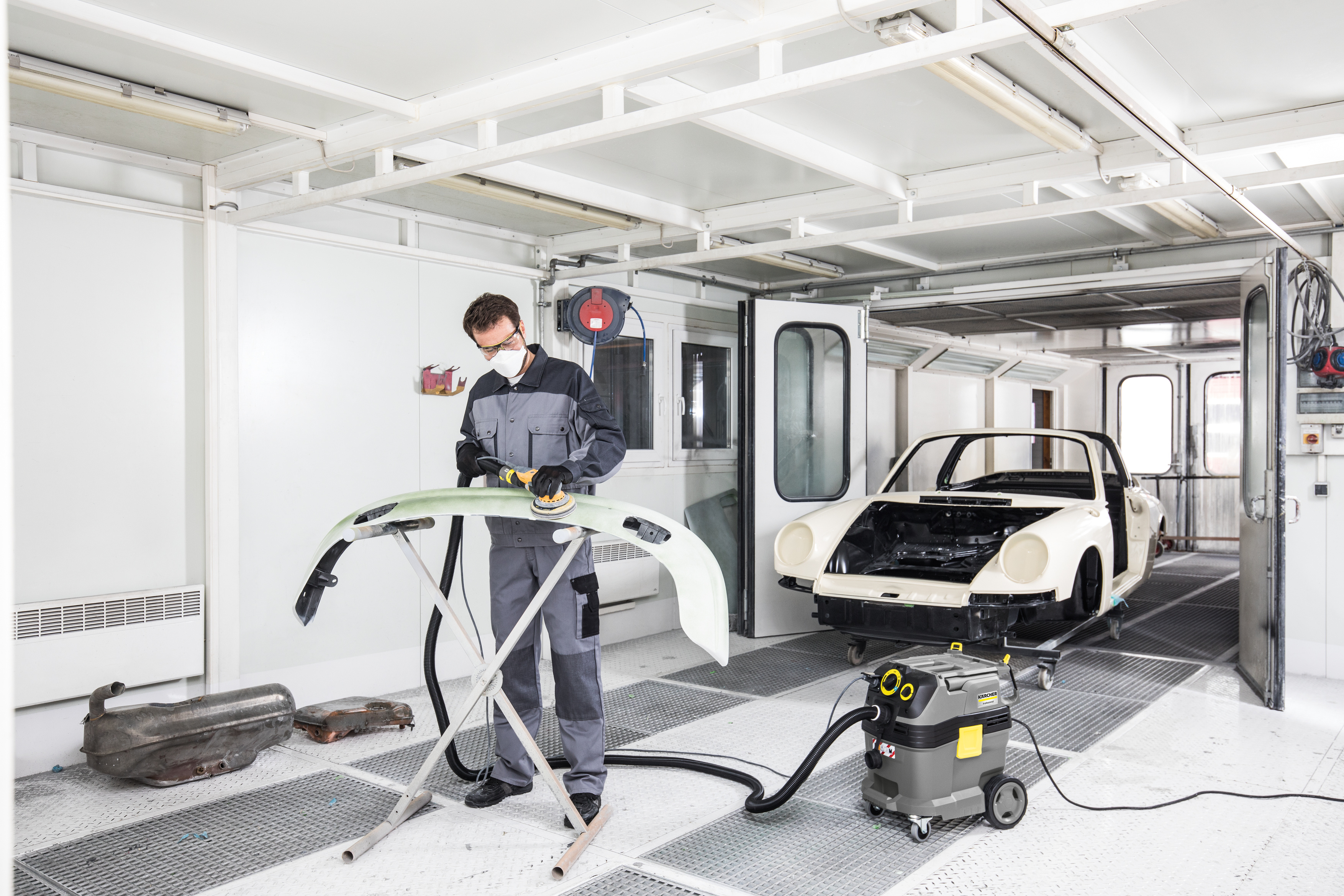 Mechaniker in Schutzkleidung schleift in heller Werkstatt eine Autostoßstange mit angeschlossenem Sauger.
