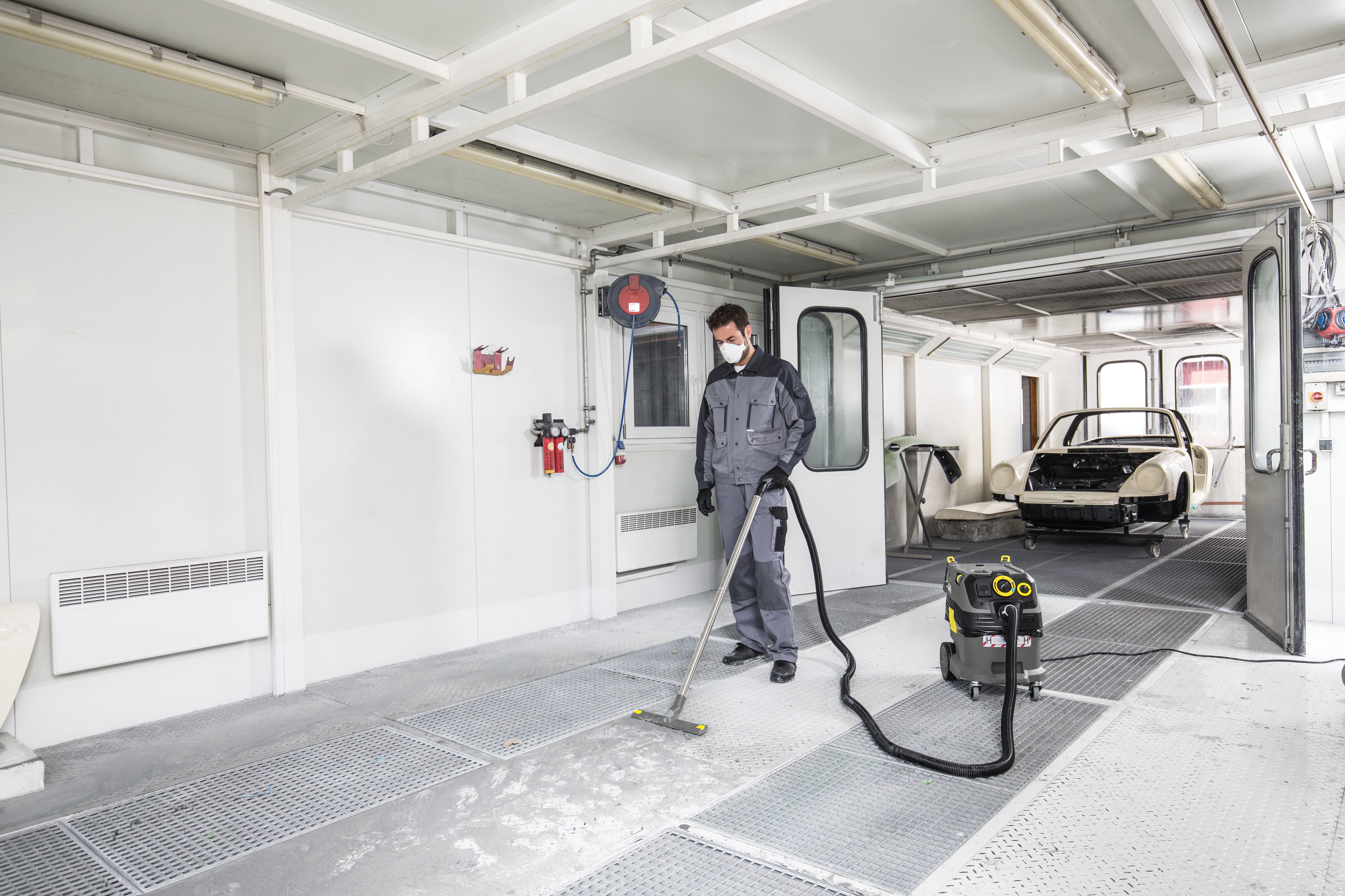 Ein Arbeiter in Schutzanzug und Maske saugt den Boden einer hellen Autowerkstatt mit einem Industriesauger.