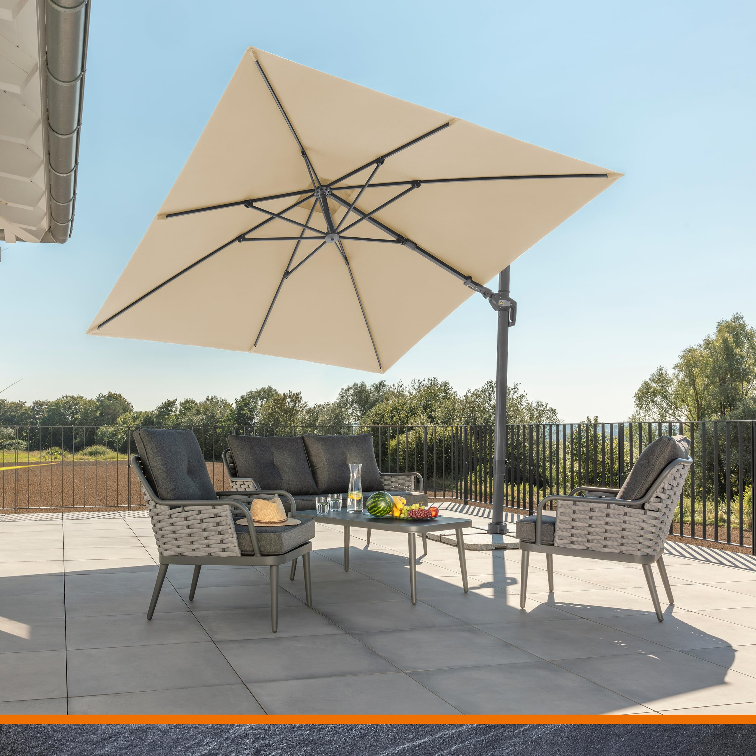 Eine graue Rattan-Sitzgruppe mit Kissen und großem beige Sonnenschirm auf einer Sonnenterrasse mit Aussicht.