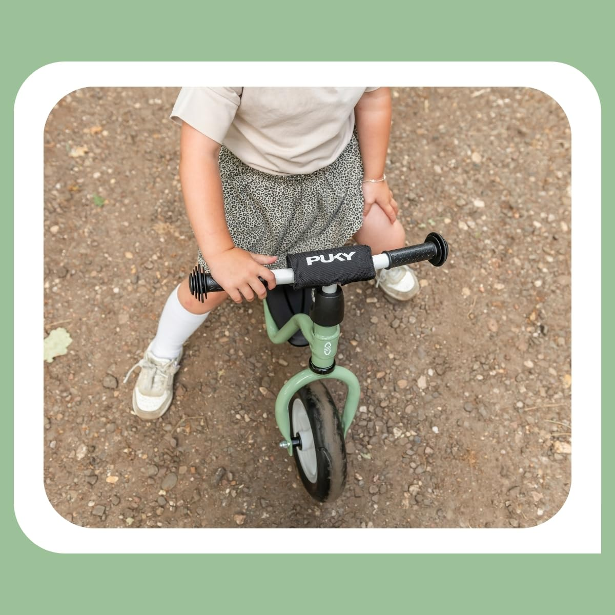 Un bambino piccolo è seduto su una bici senza pedali Puky verde su un sentiero di ghiaia.