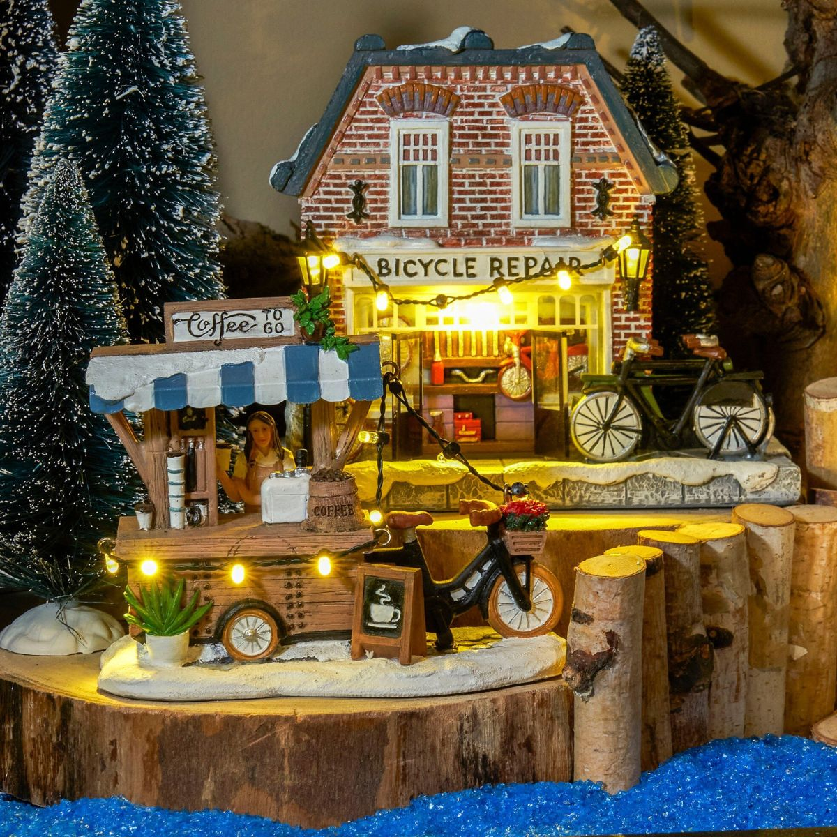Een miniatuur winterdorp met verlichte koffiekar, fietsenmaker, besneeuwde bomen en houten hek.