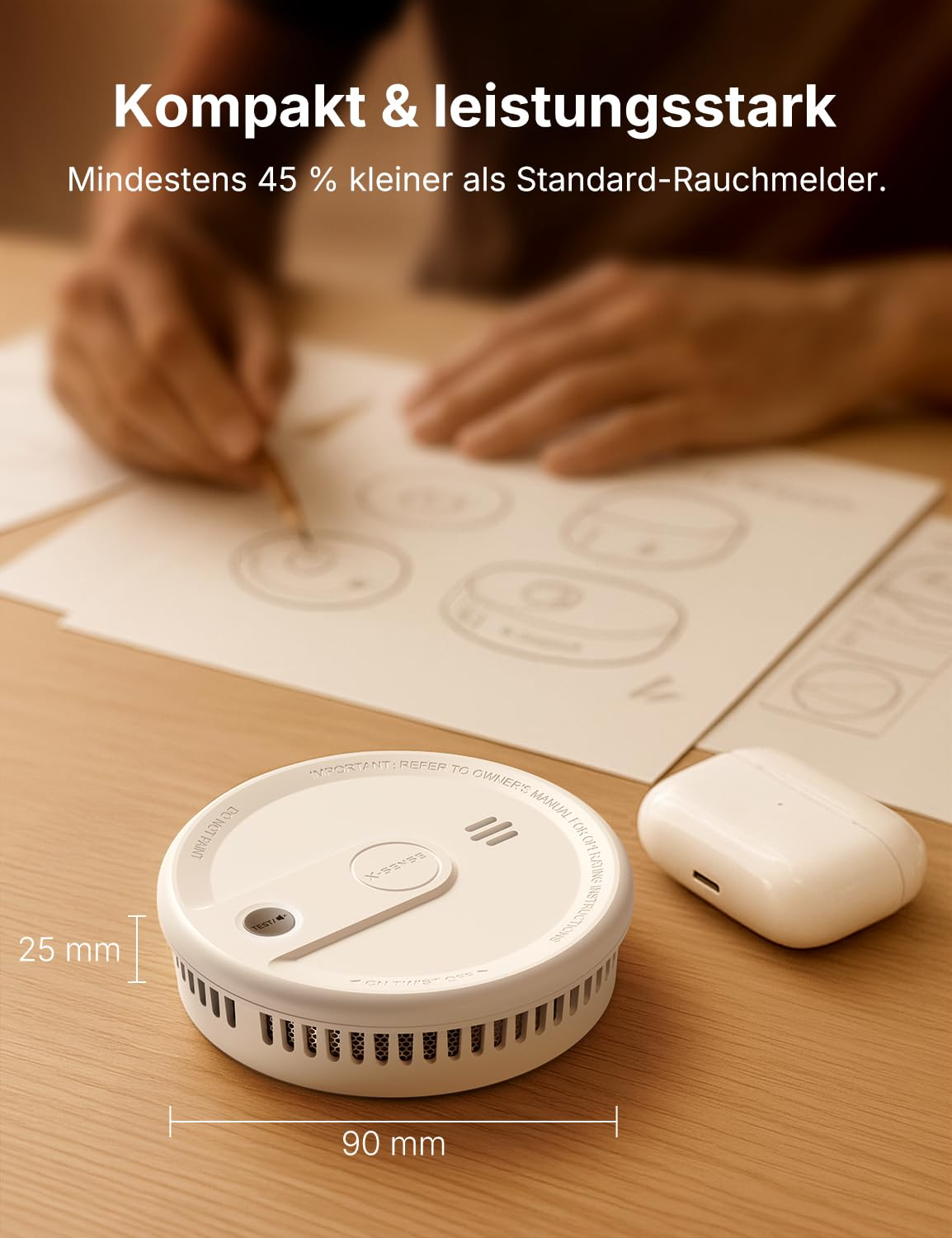 Ein kleiner weißer Rauchmelder auf einem Holztisch mit Designskizzen im Hintergrund und kabellosen Ohrhörern daneben.