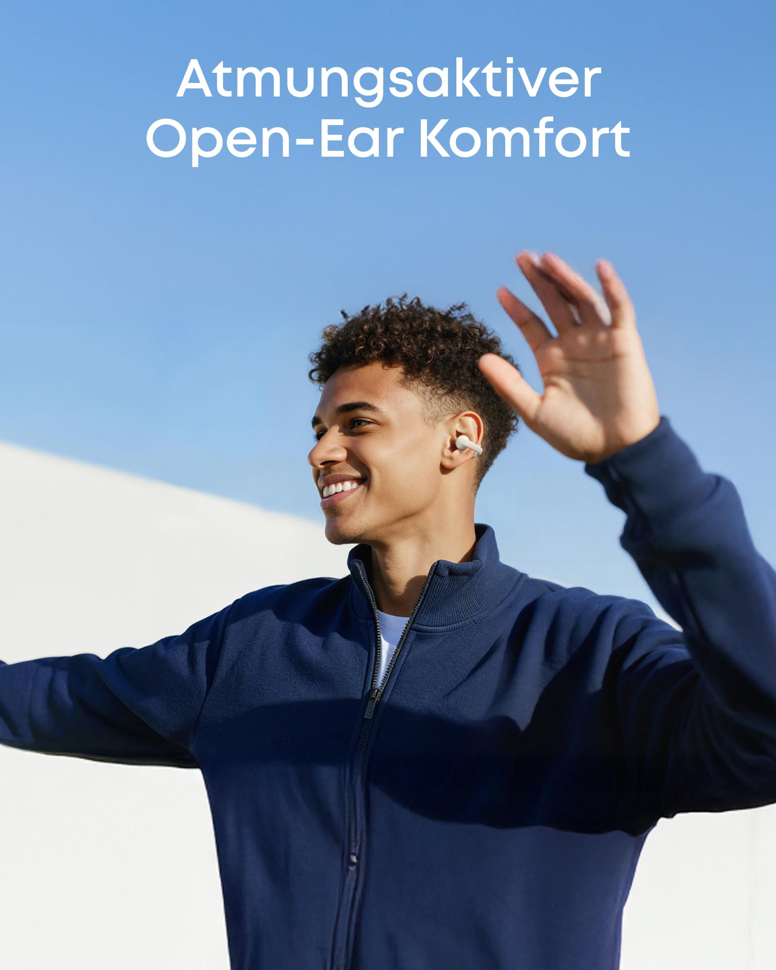Ein lächelnder junger Mann mit Kopfhörern hebt im Freien winkend die Hand unter blauem Himmel.