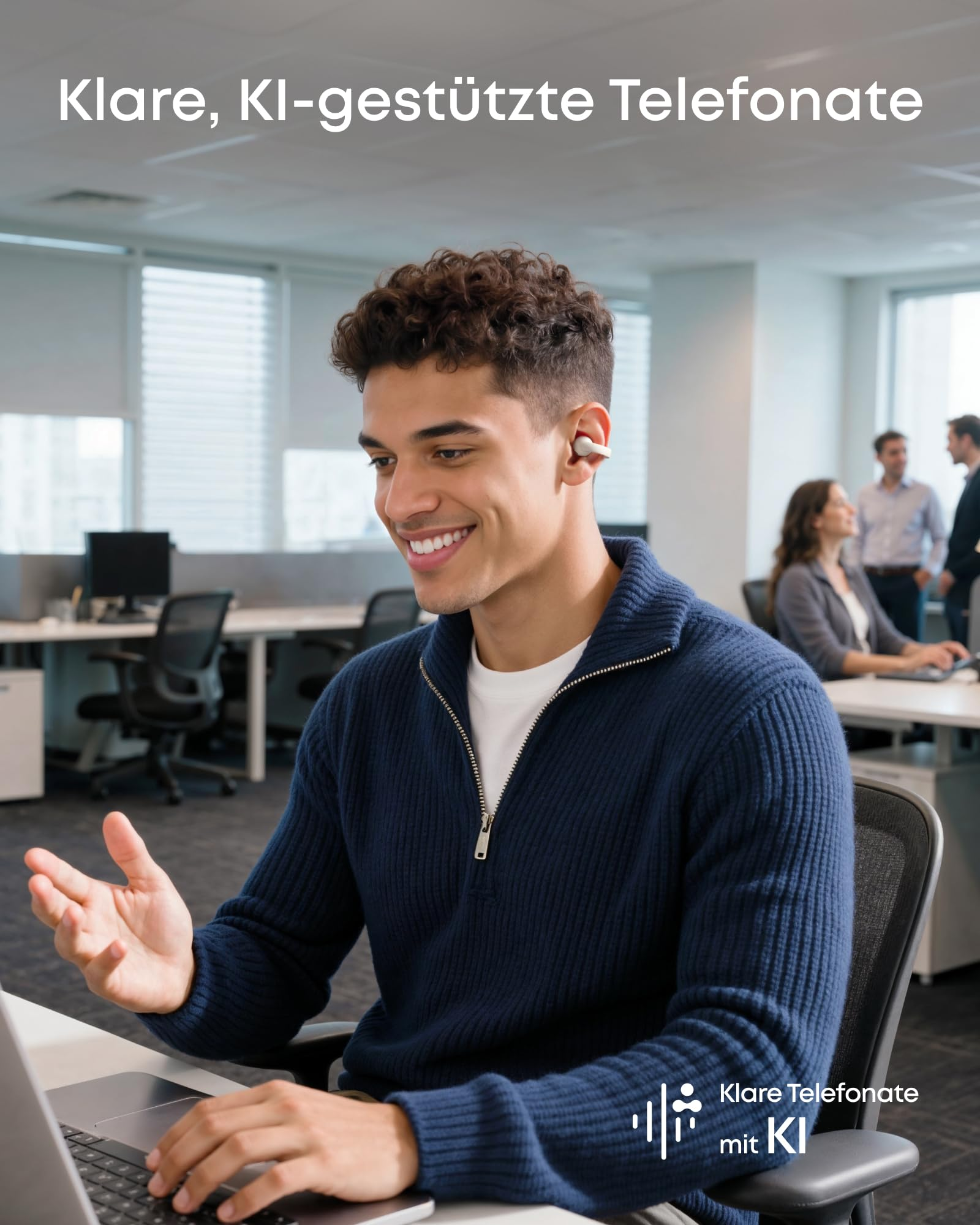 Ein junger Mann mit dunklen Haaren und blauem Pullover sitzt lächelnd im Büro an einem Laptop und telefoniert.