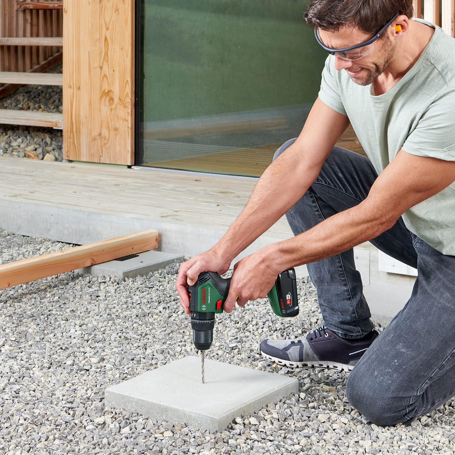 Ein Mann kniet auf Kies und bohrt mit einem Akkubohrer in eine Betonplatte vor einem Haus.