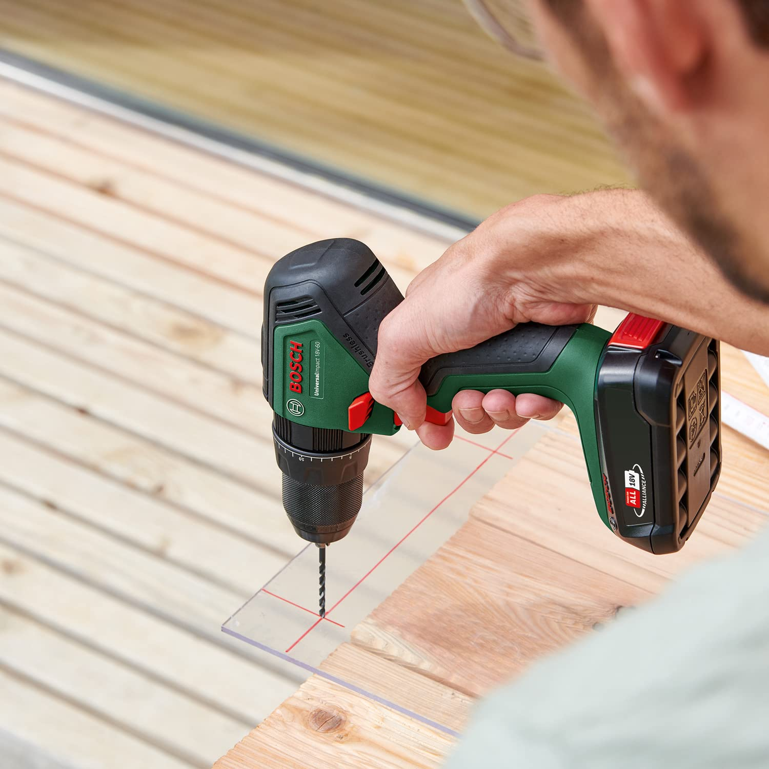 Ein Handwerker bohrt mit einer grünen Bosch-Akkubohrmaschine ein Loch in eine transparente Platte auf Holz.
