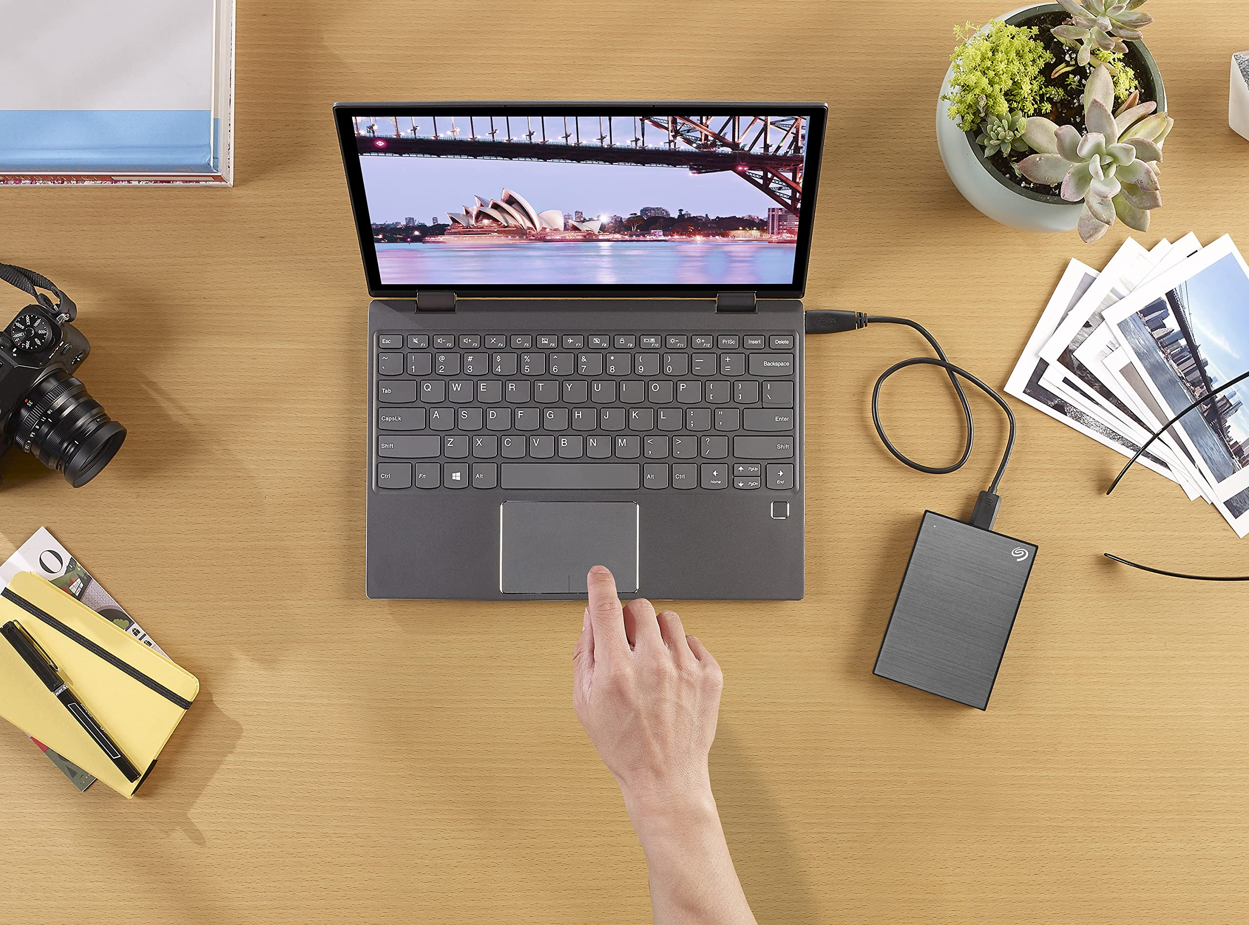 Eine Hand bedient einen Laptop auf einem Holztisch mit Kamera, externer Festplatte, Fotos, Pflanzen und Notizbuch.