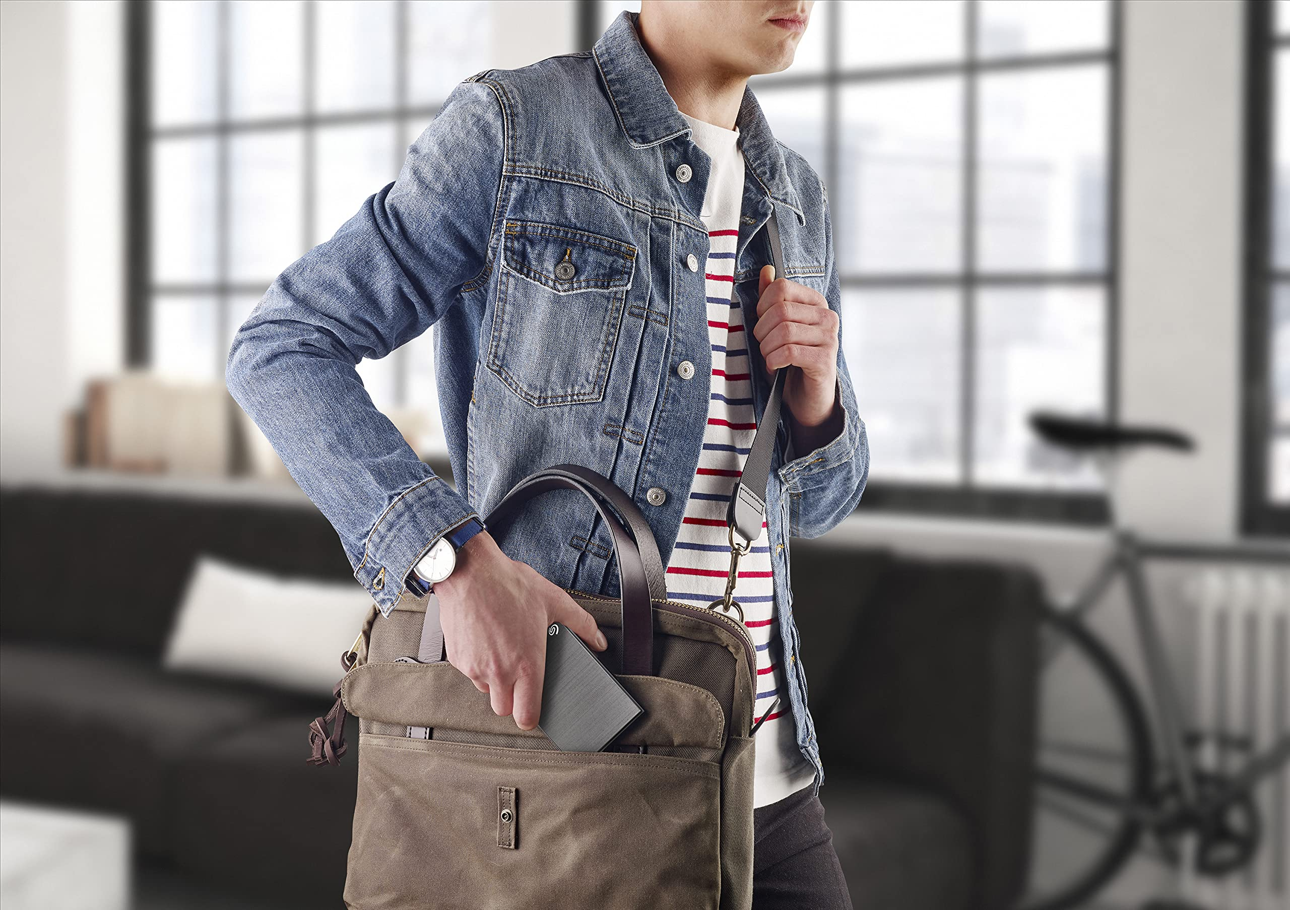 Ein Mann im Jeanshemd und gestreiftem Shirt legt ein Gadget in eine braune Tasche.