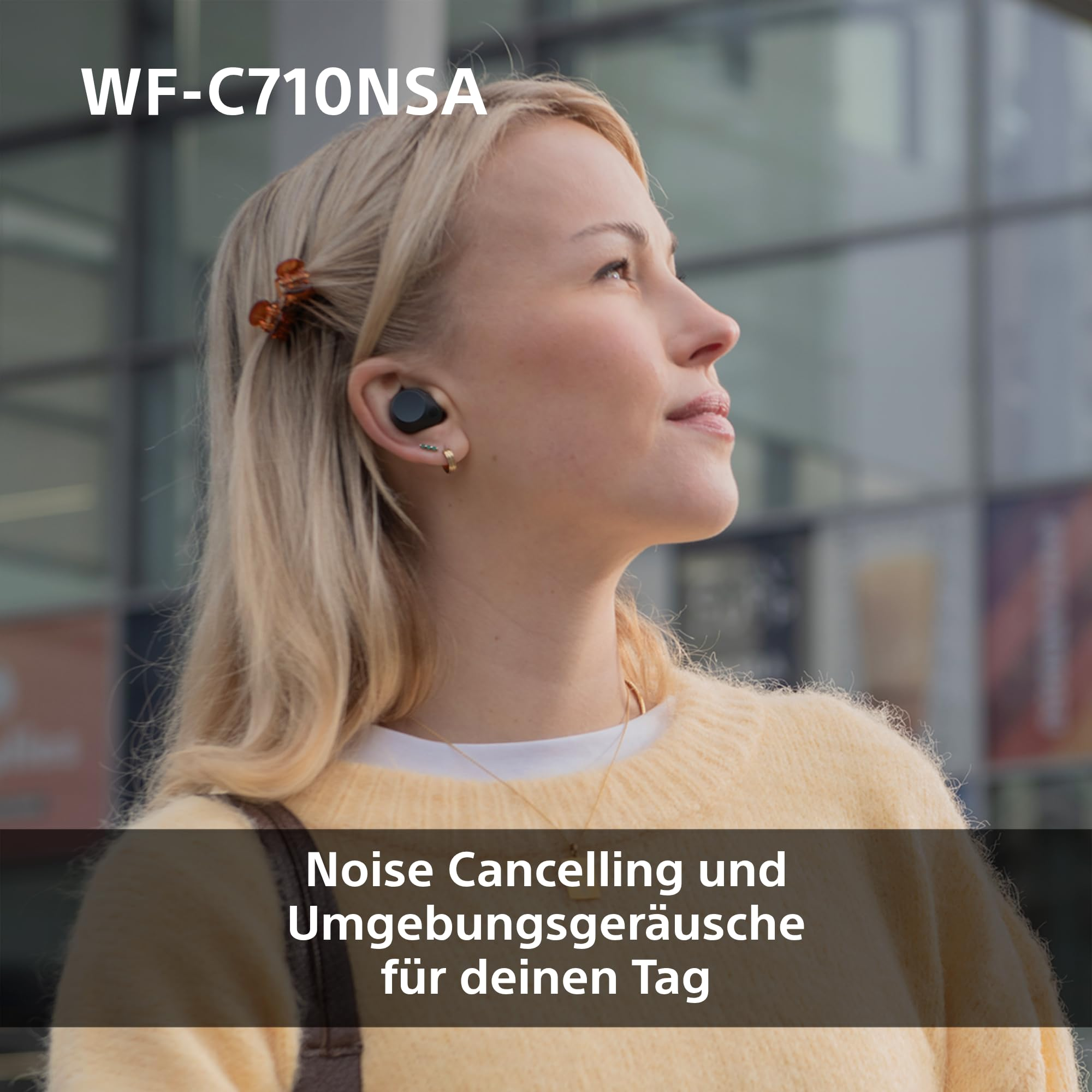 Una donna bionda indossa auricolari neri e guarda in alto davanti a un edificio moderno.