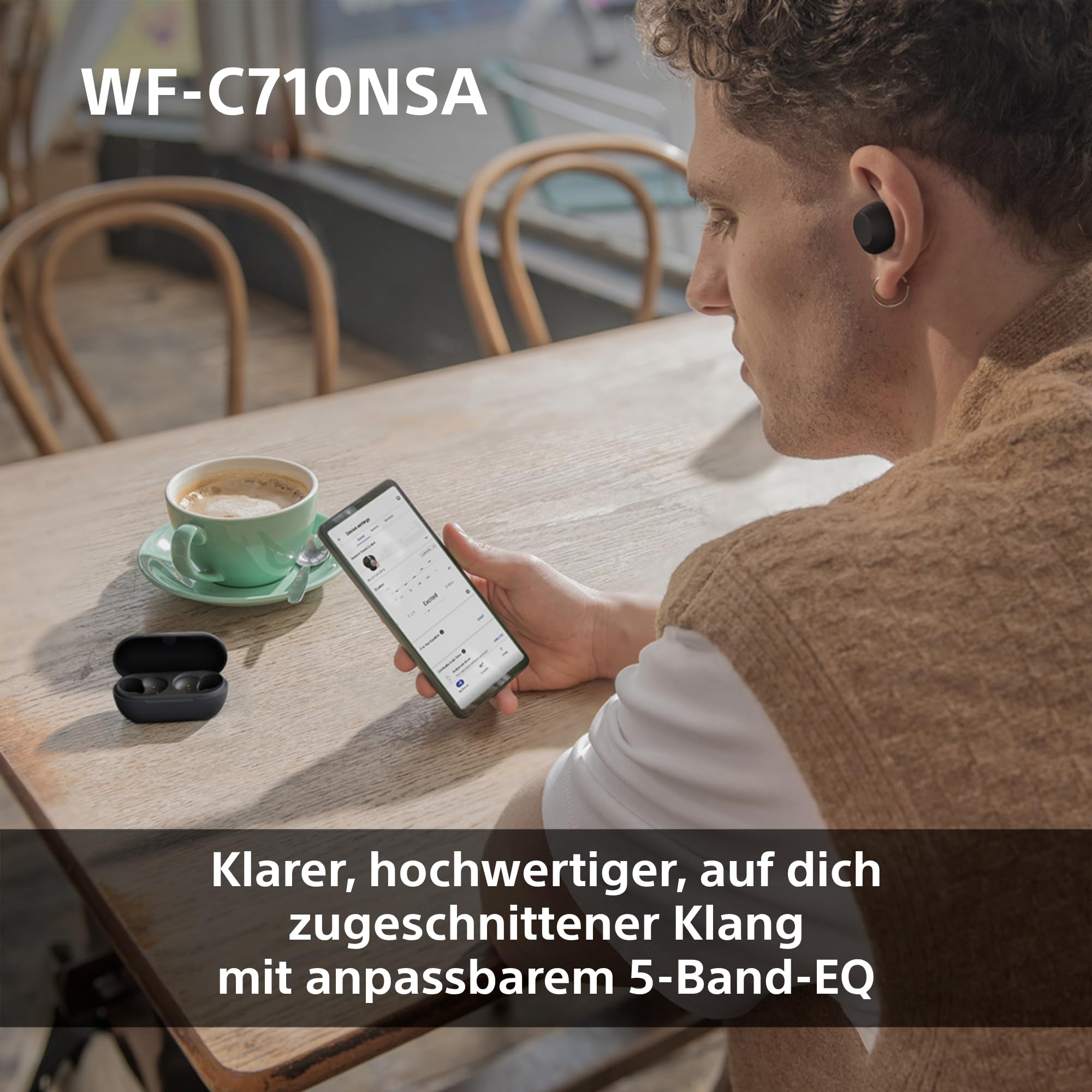 Un uomo con auricolari siede a un tavolo di legno, tenendo uno smartphone e guardando una tazza di caffè.