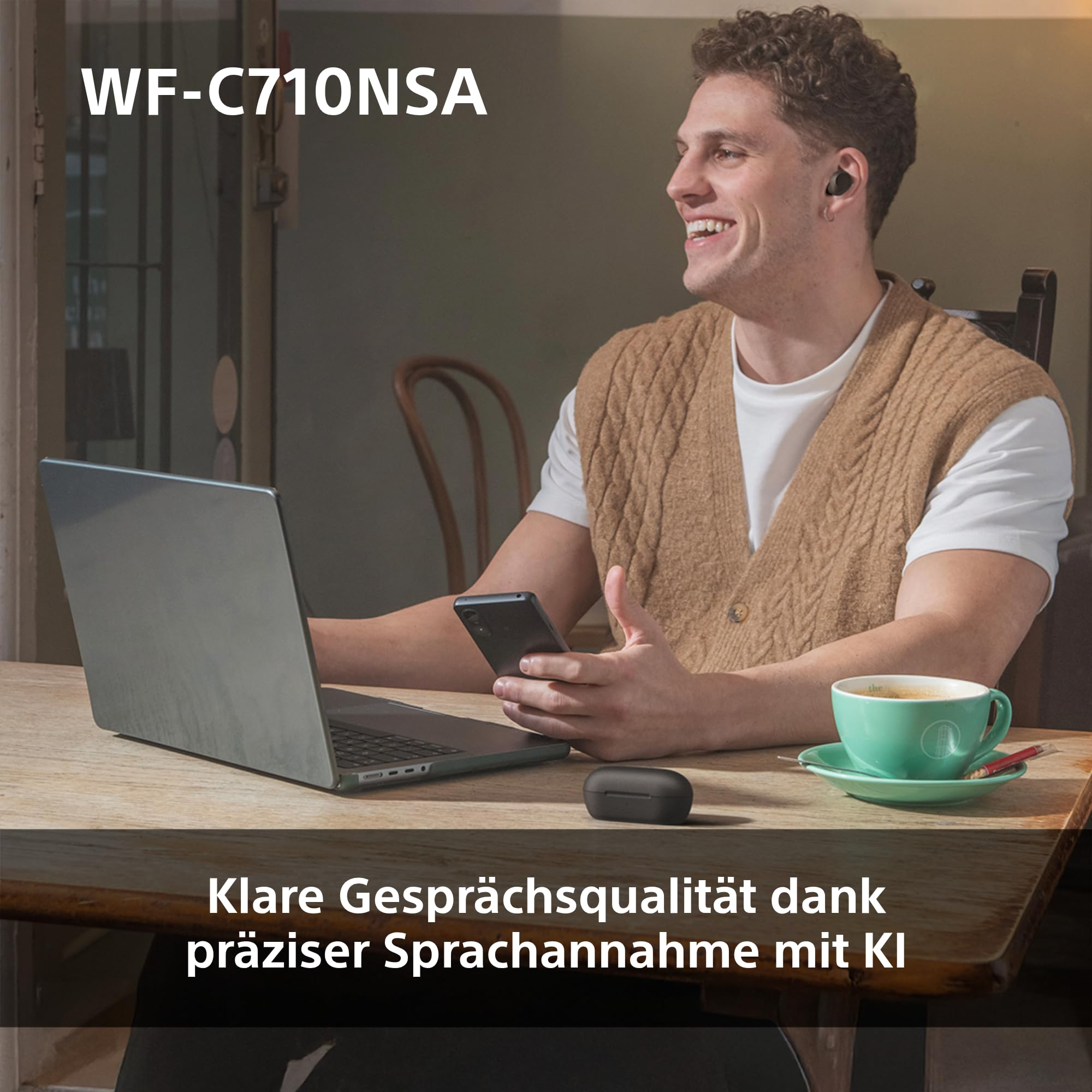 Un uomo sorridente con auricolari wireless è seduto a un tavolo di legno con un laptop, uno smartphone e una tazza di caffè.