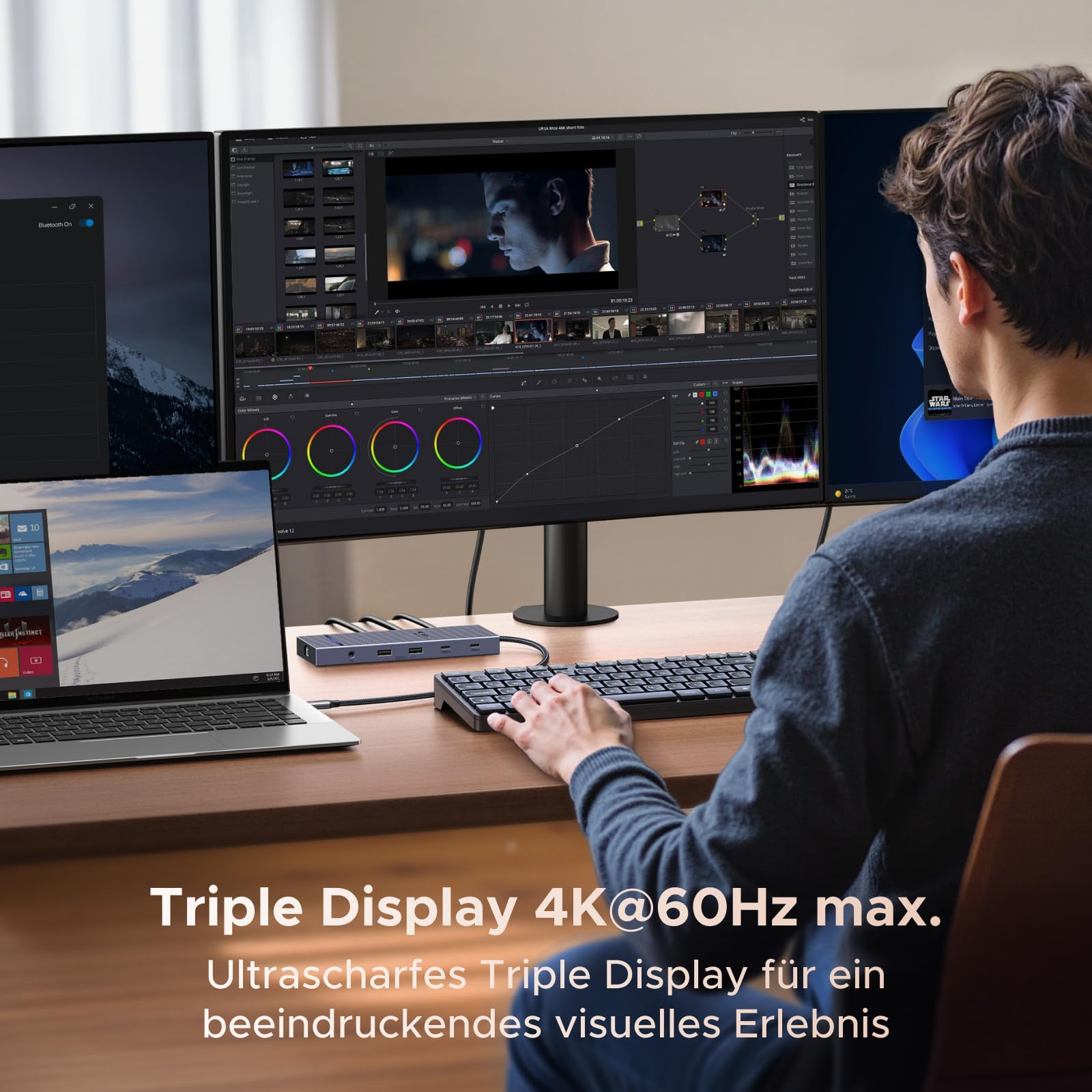 Un uomo siede a una scrivania di legno con laptop, tastiera e tre monitor collegati per video editing.