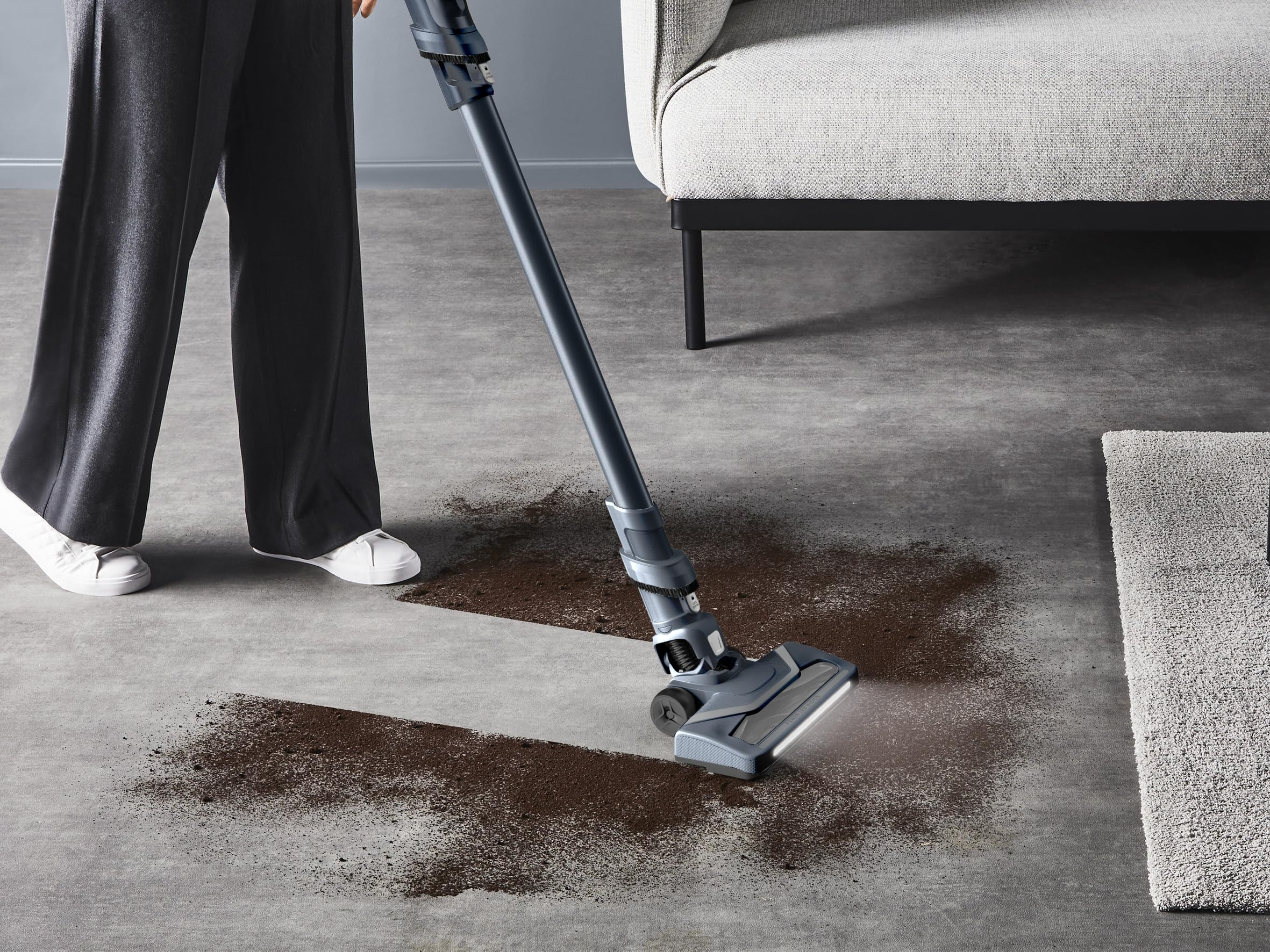 Persona aspira sporco marrone su pavimento in cemento grigio con un aspirapolvere blu.