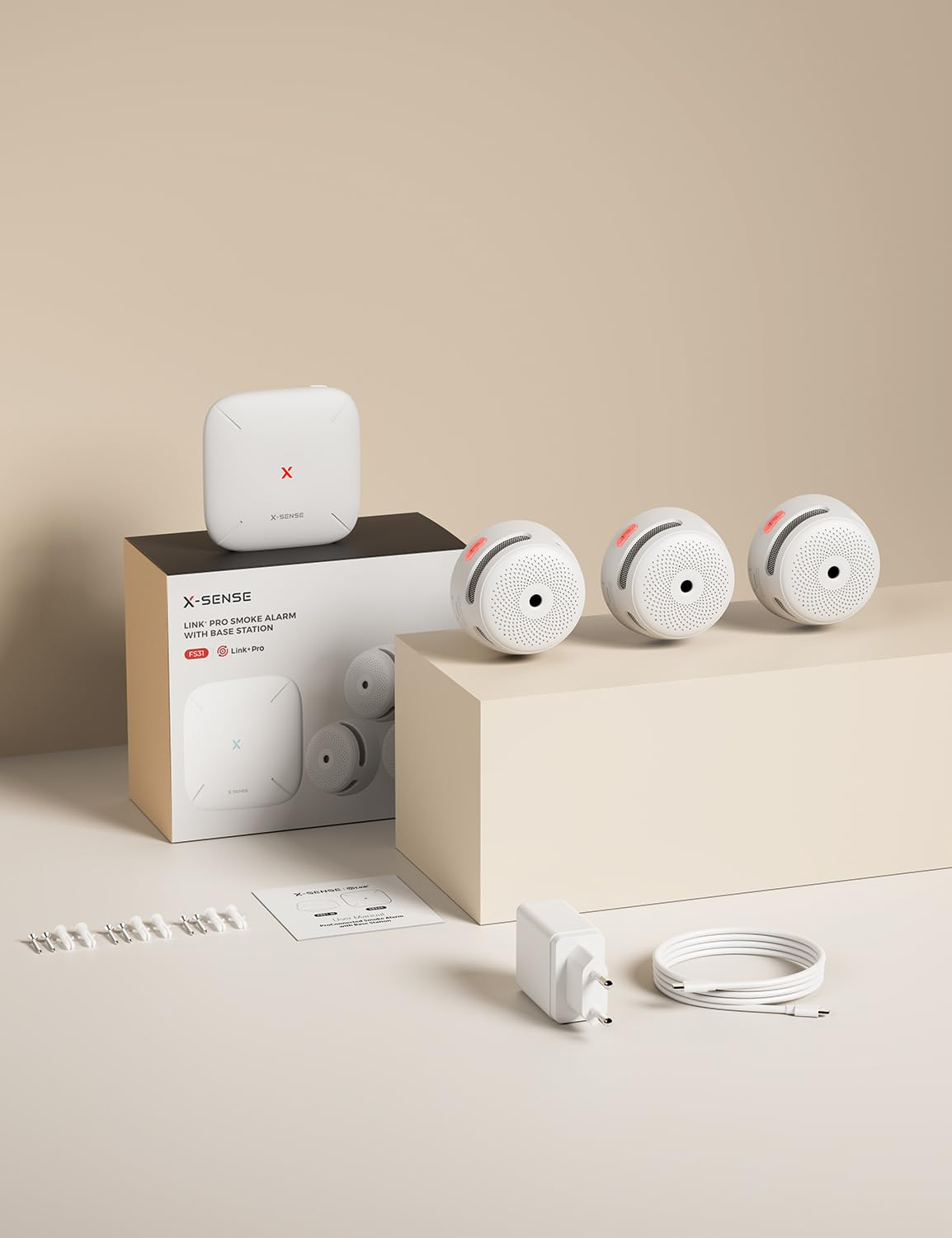 Kit rilevatore di fumo X-Sense con base, tre allarmi, alimentatore, cavo e viti su un tavolo beige.