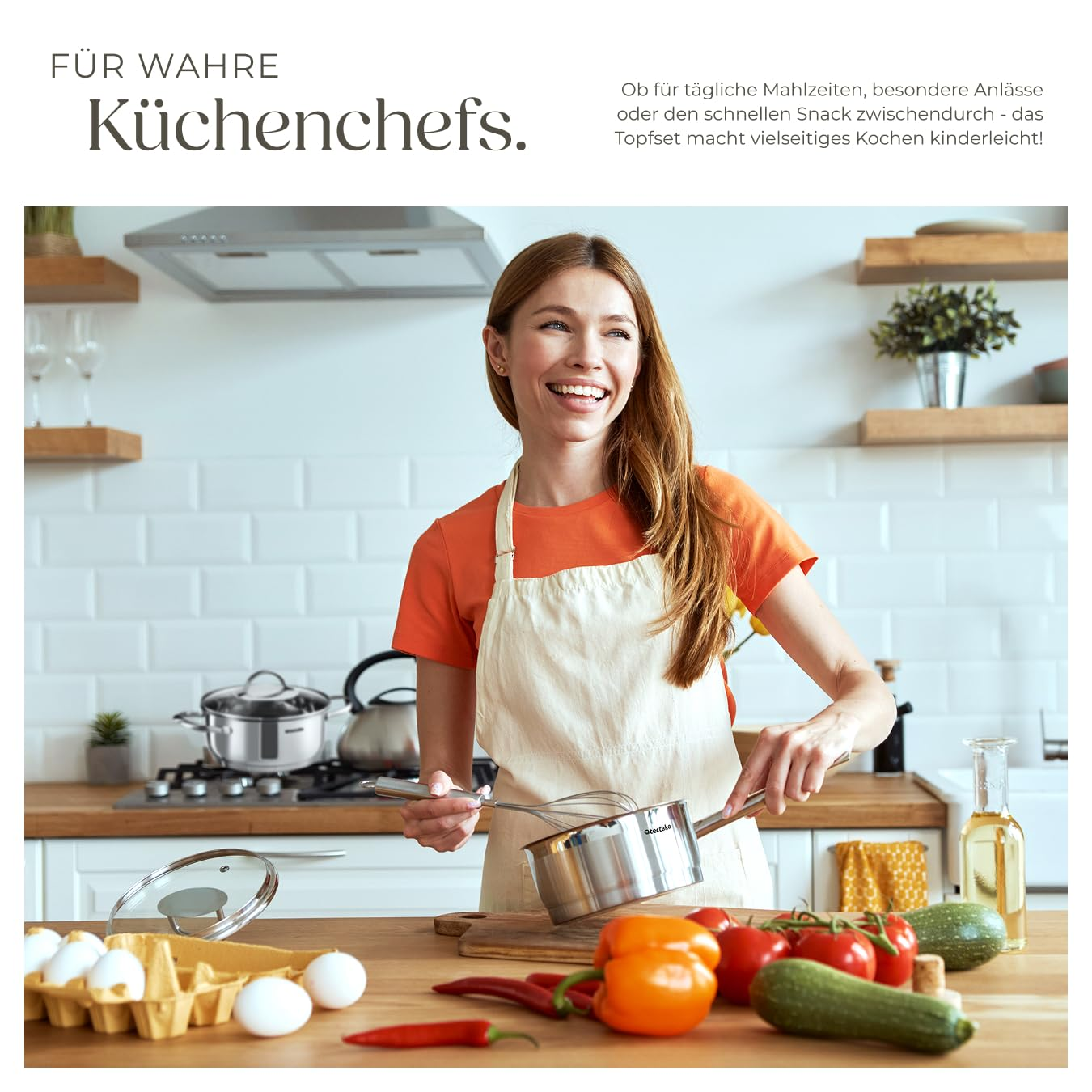 Donna sorridente in cucina con frusta e pentola, verdure e uova sul bancone.