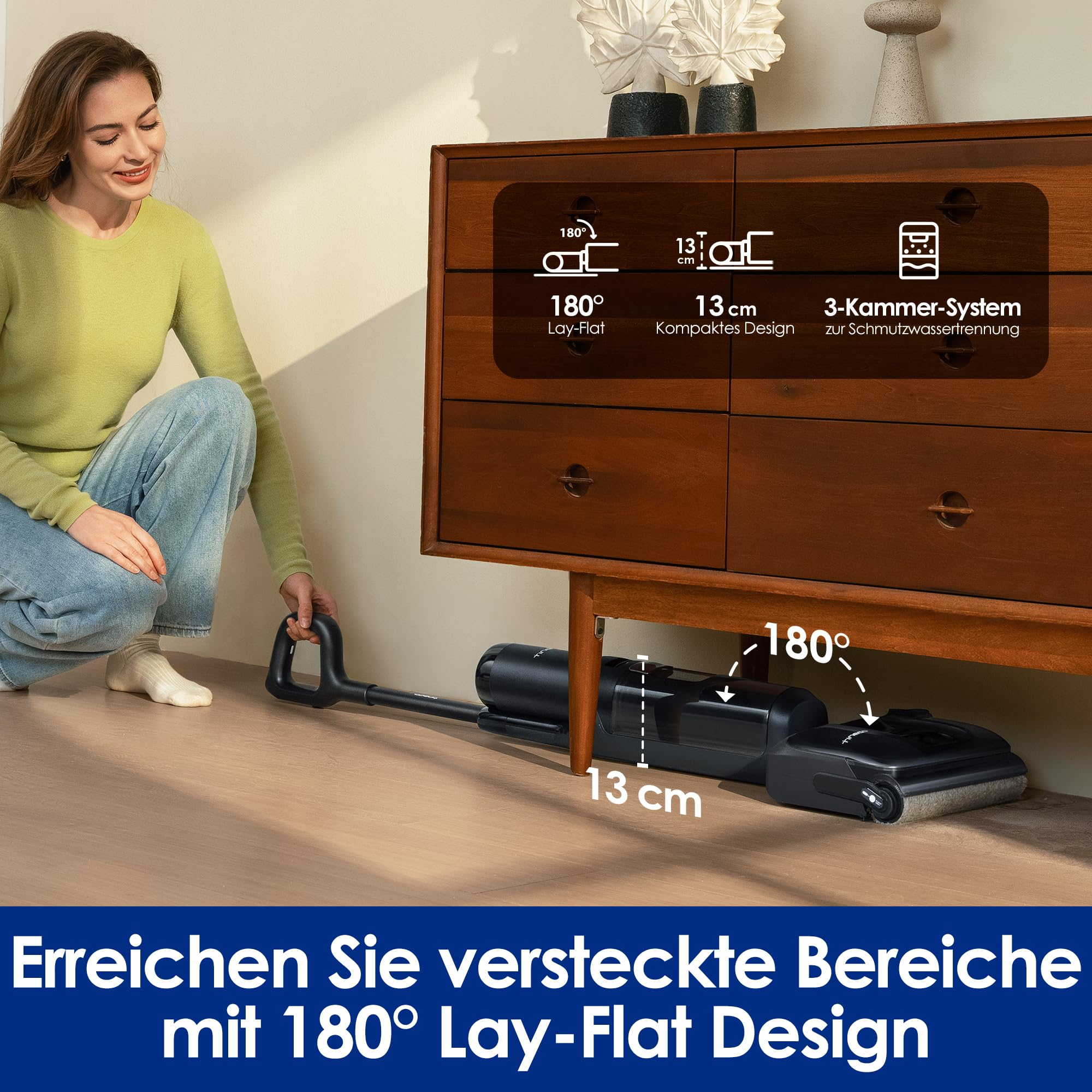 Eine Frau kniet neben einem flachen Staubsauger, der unter einer Holzkommode putzt.