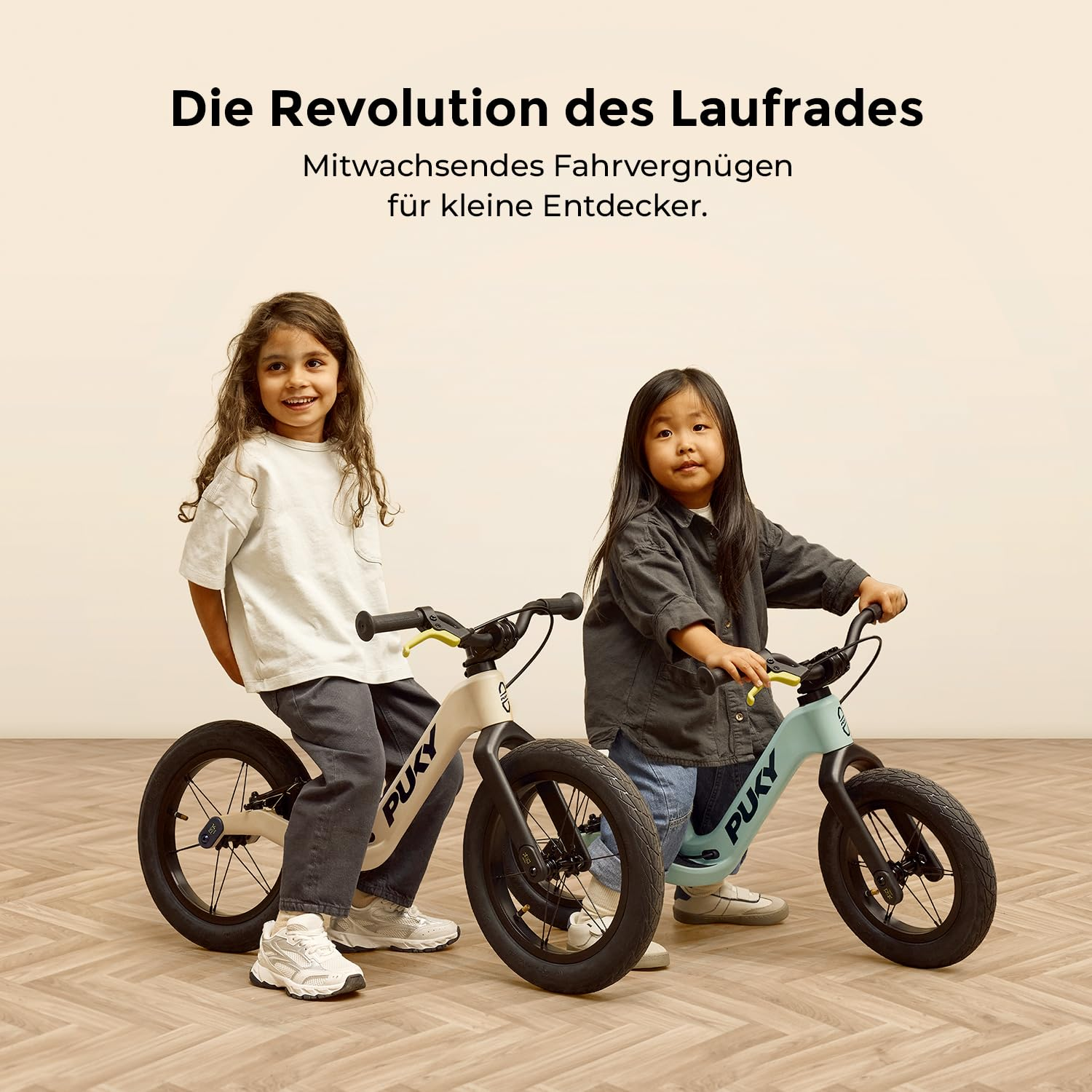 Zwei Kinder lächeln auf Puky-Laufrädern auf einem Holzfußboden, mit deutschem Text oben.