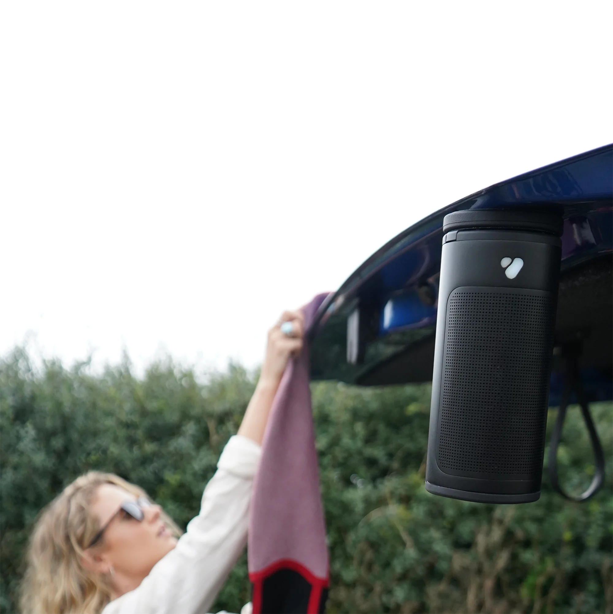 Un haut-parleur noir est suspendu à une portière de voiture bleue ouverte et une femme tient une serviette.