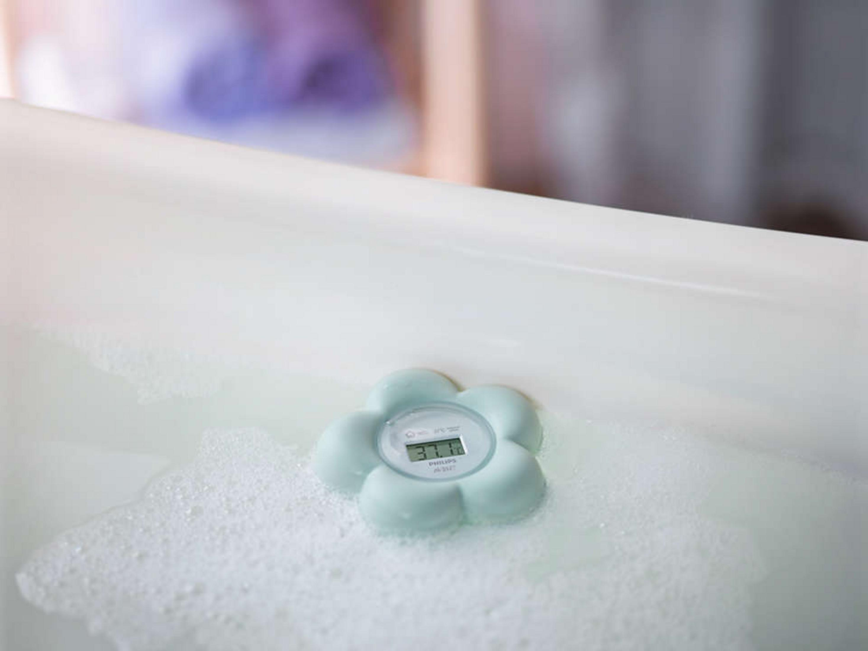 Ein mintgrünes, blumenförmiges Badethermometer schwimmt im Badewasser mit Schaum.