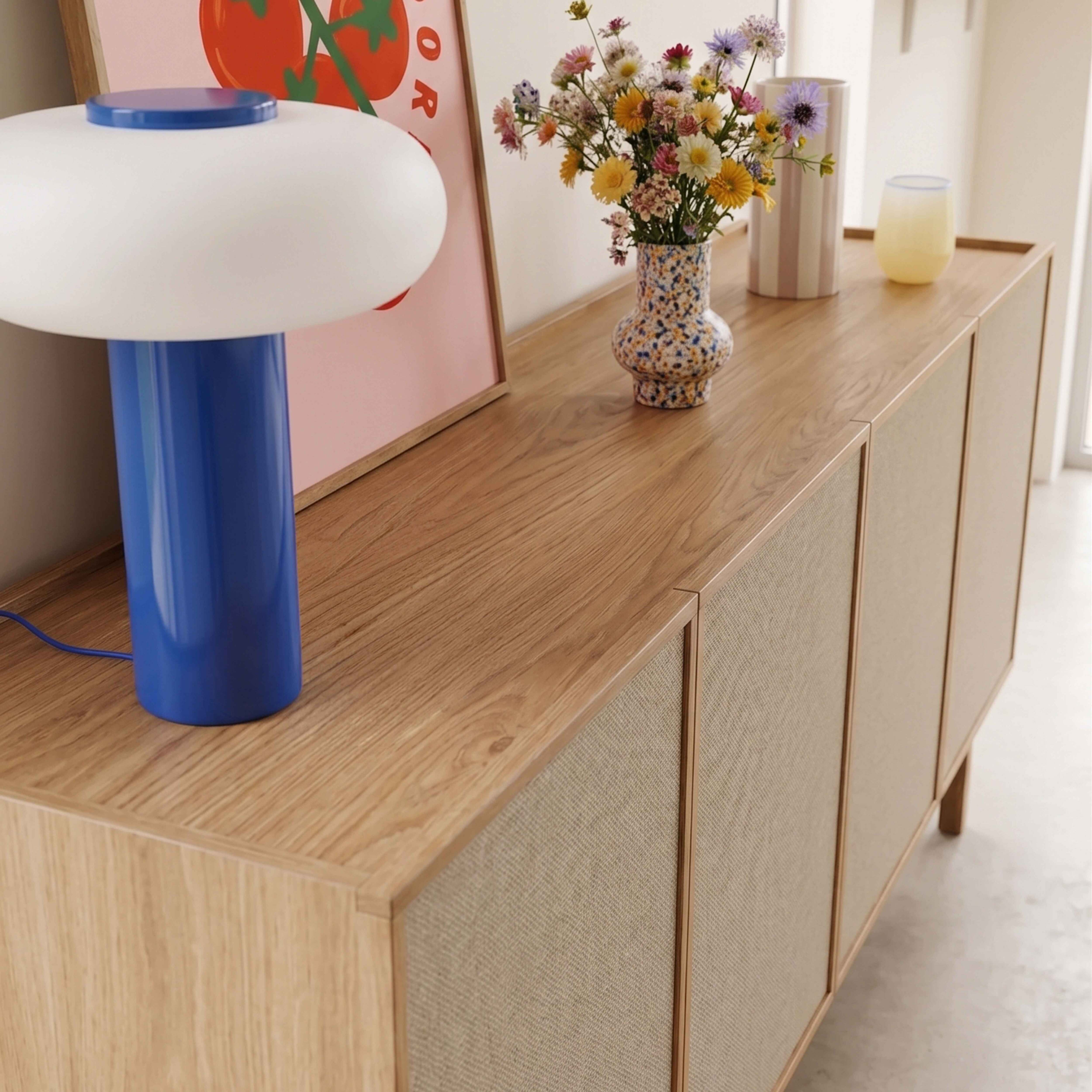 Holzsideboard mit blauer Tischlampe, bunter Blumenvase, eingerahmtem Poster und Deko auf heller Wand.