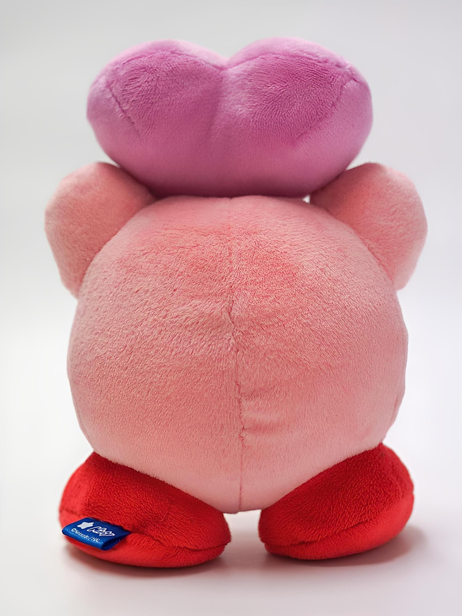 Vista trasera de un personaje de peluche rosa redondo con pies rojos y un gran corazón rosa en la cabeza.